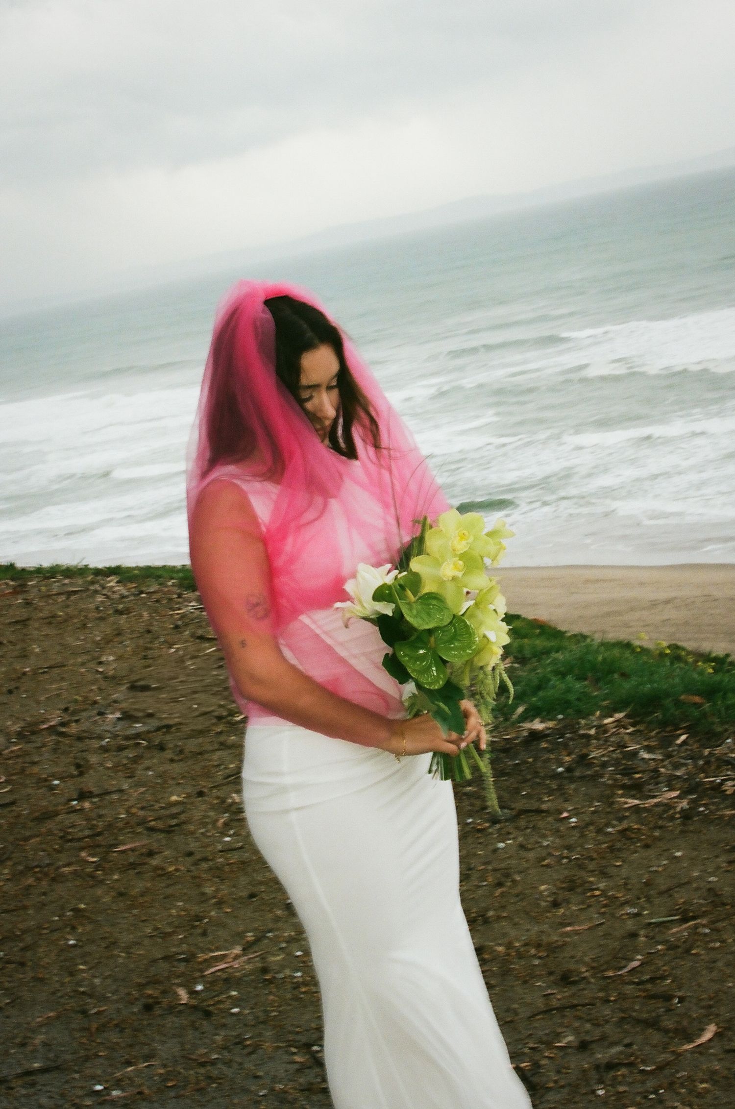 Bride in white gown and pink veil holding green bouquet on a coastal cliff overlooking the ocean.
