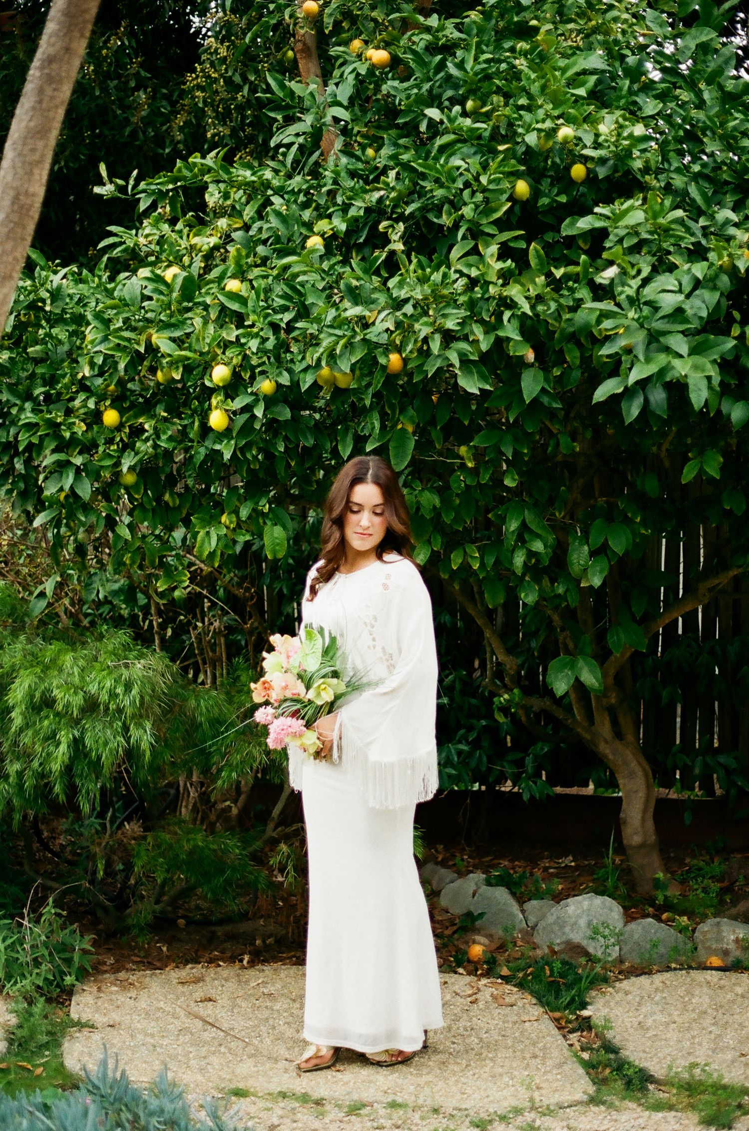 Bride in white fringe top and skirt standing beneath a lemon tree holding a soft pastel bouquet.