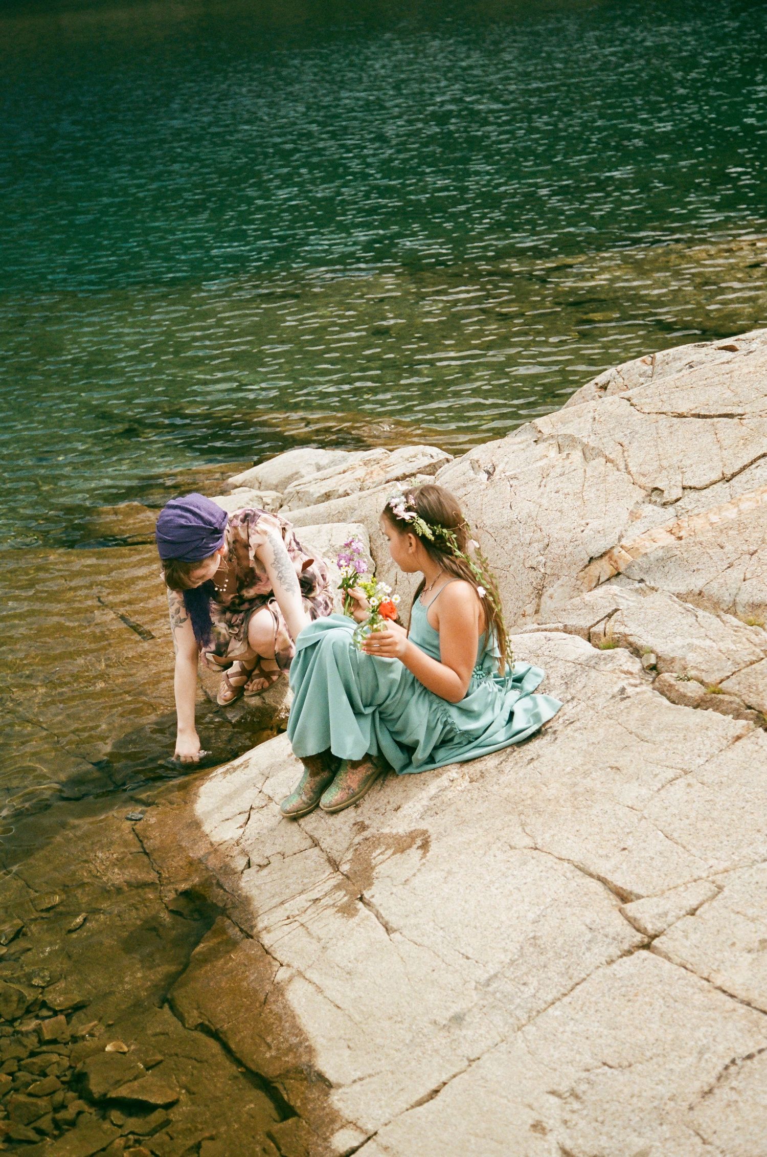 Two young women in floral crowns and green dresses sit on rocky shore by turquoise water, sharing a bouquet of flowers.