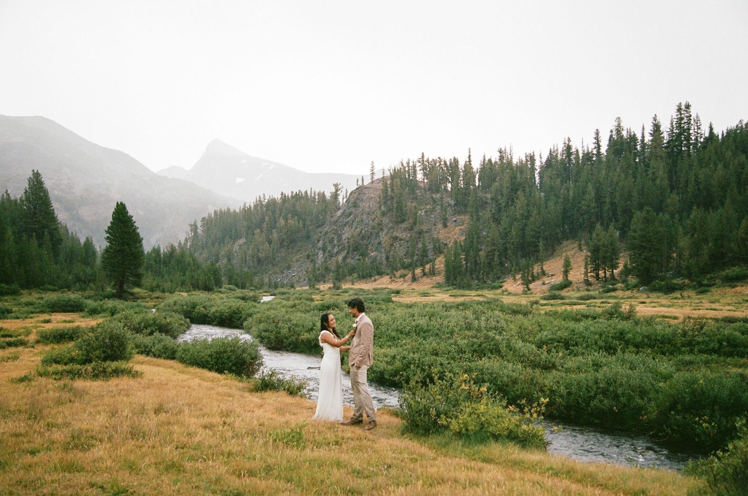 Couple shares a romantic dance in a lush mountain meadow beside a winding stream, surrounded by evergreen forests.