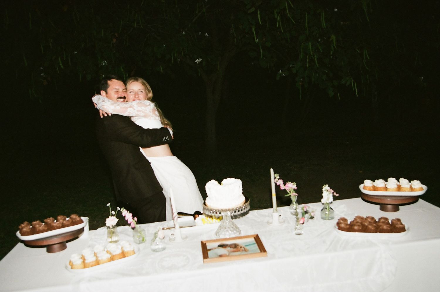 Bride and groom embracing behind a wedding dessert table with cake and treats at an outdoor evening reception.