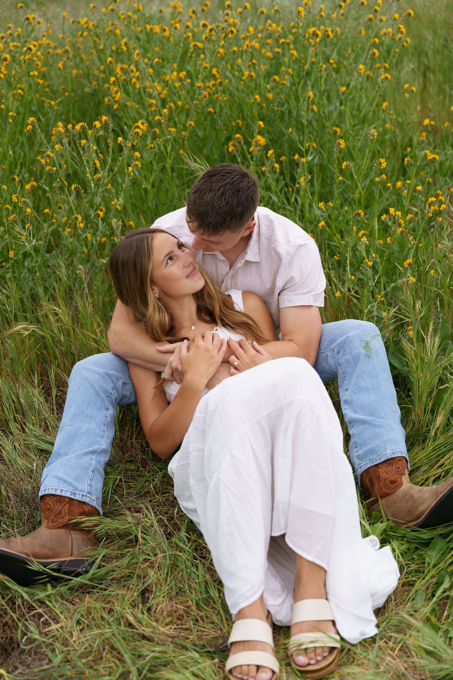 Couple sits intimately in green meadow with yellow wildflowers, man kissing womans cheek tenderly.