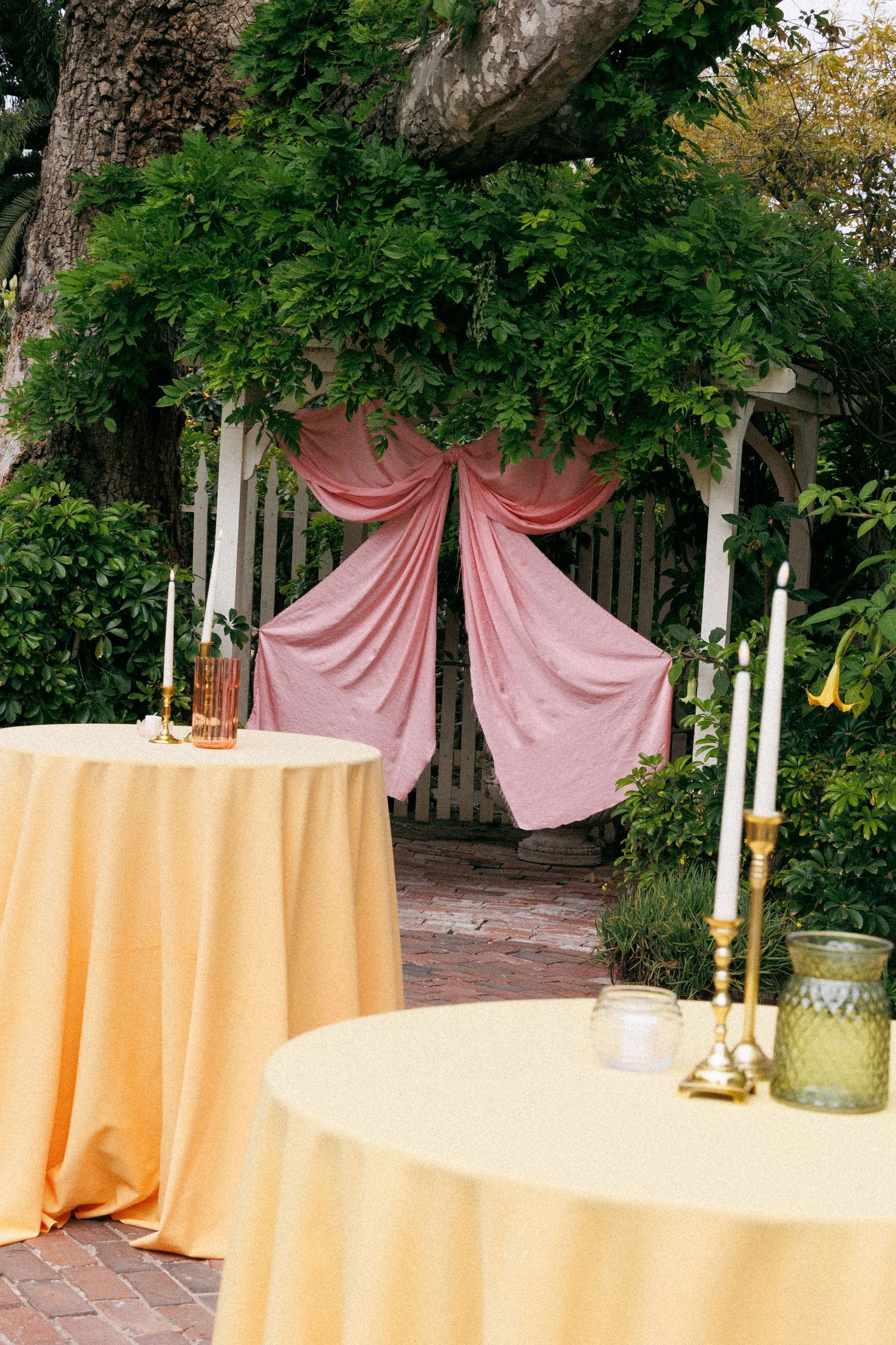 Outdoor wedding reception setup with yellow linen tables and pink draped arbor amid lush greenery.