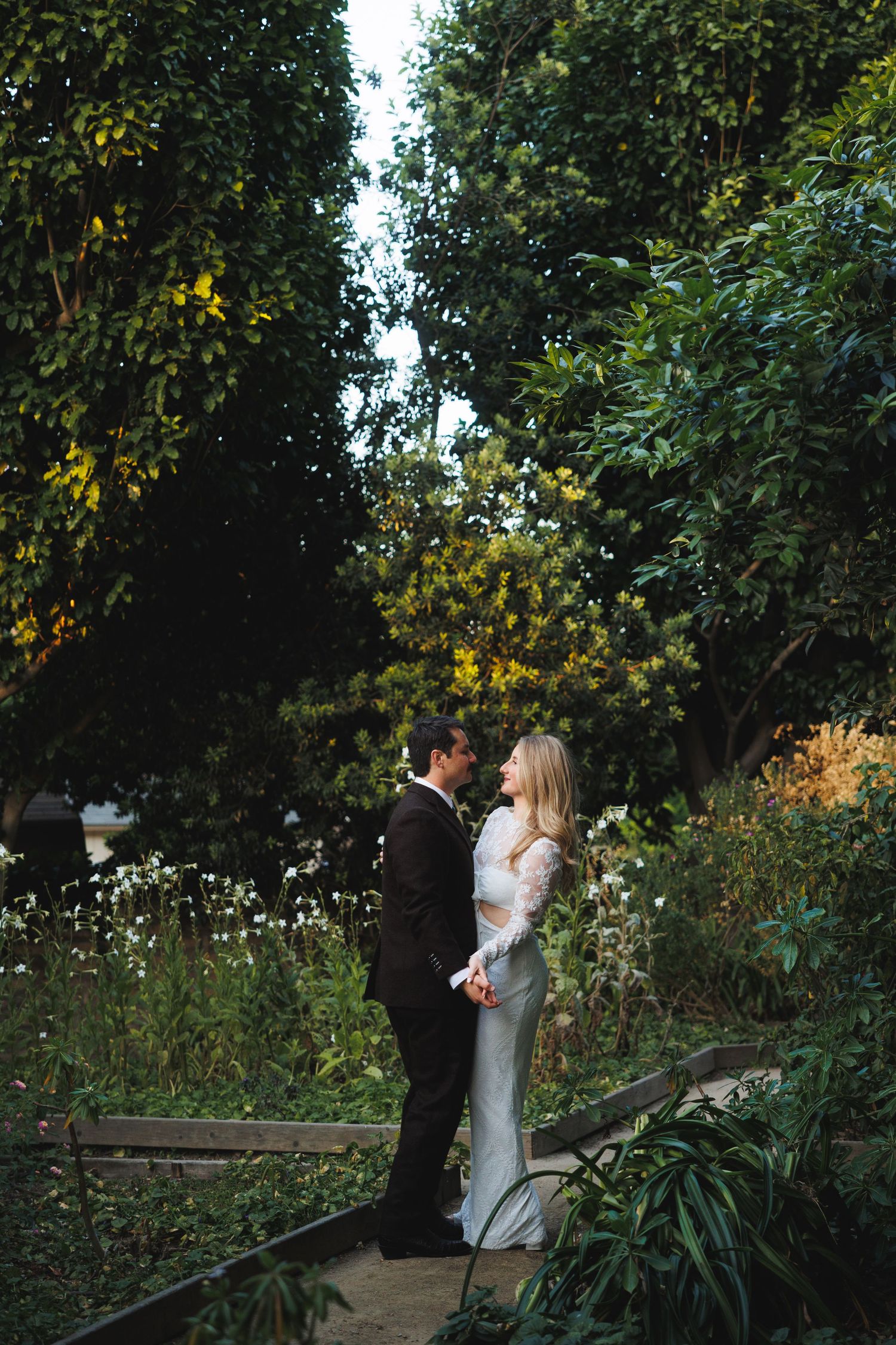 Couple in formal attire share a romantic moment on a garden path surrounded by lush green trees.