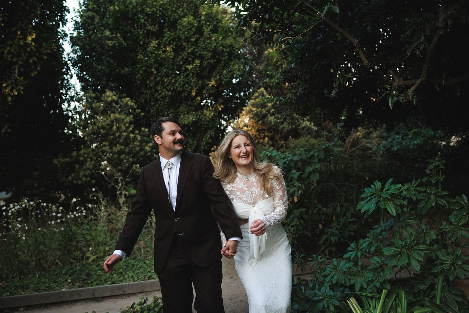 Happy bride and groom walking hand in hand through lush garden greenery on their wedding day.