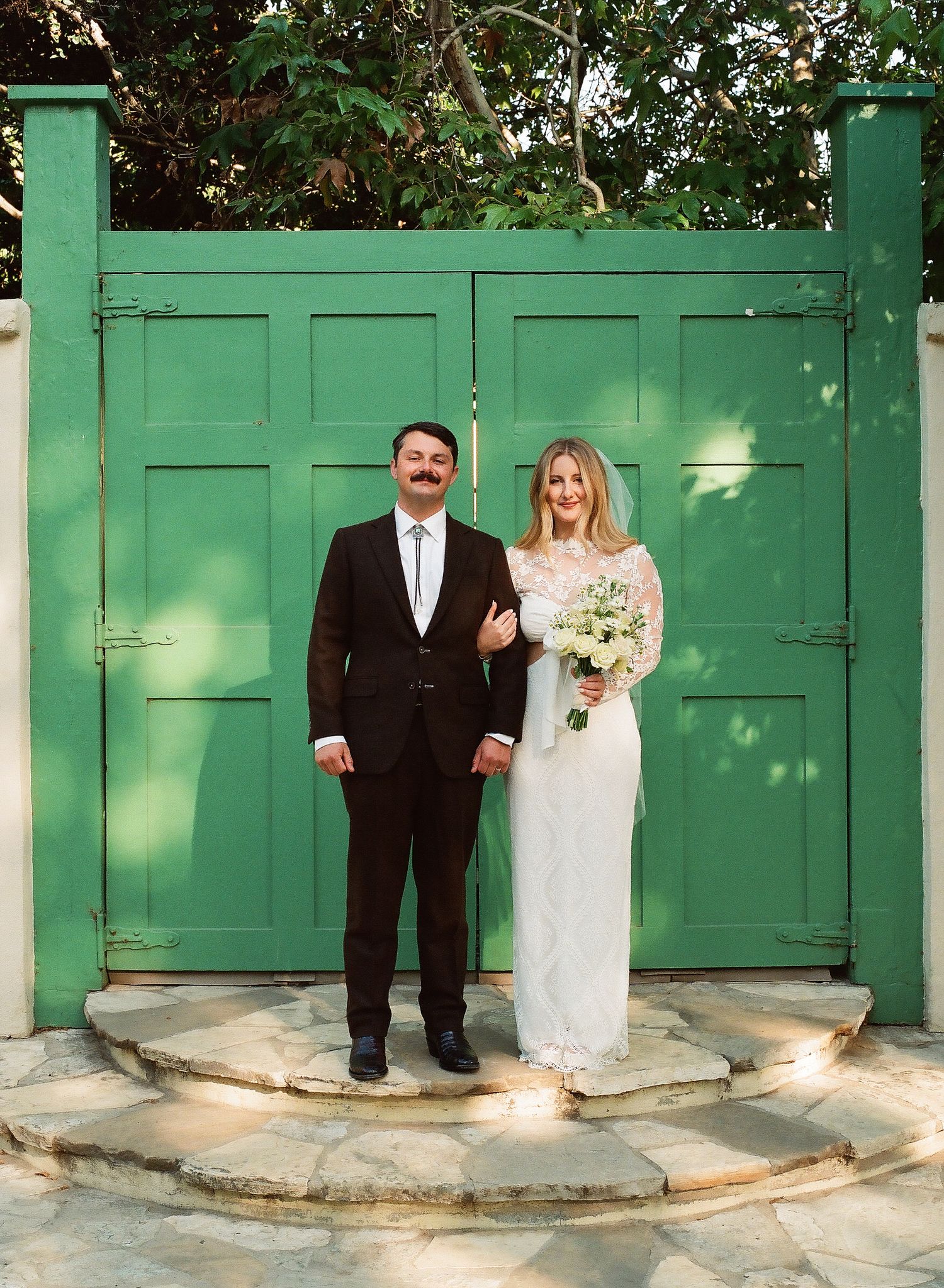 Bride and groom posing in front of green wooden gates, bride holding white bouquet, groom in black suit with white tie.