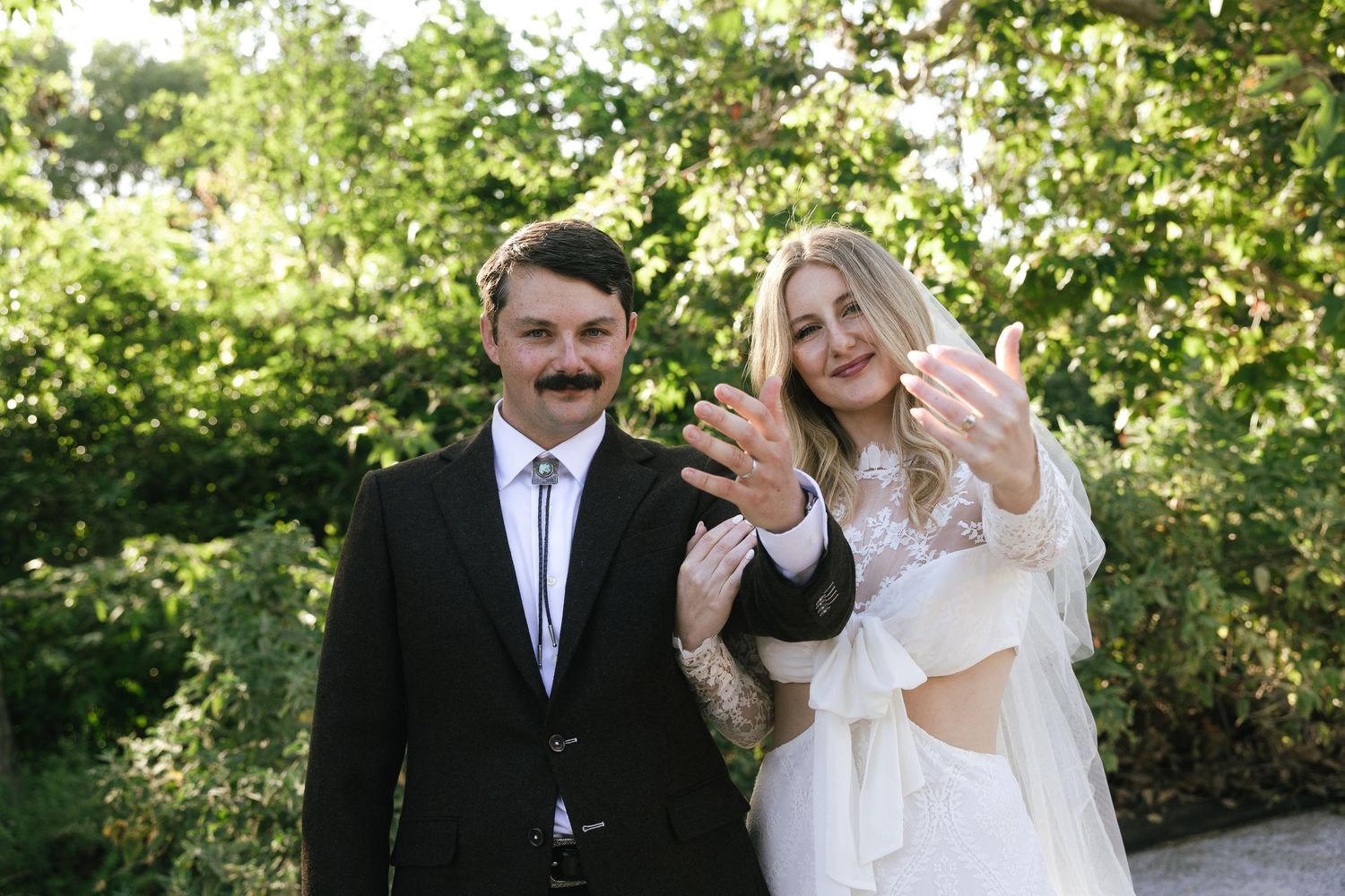 Newlywed couple in wedding attire showing off rings outdoors, groom in black suit with mustache, bride in white dress.