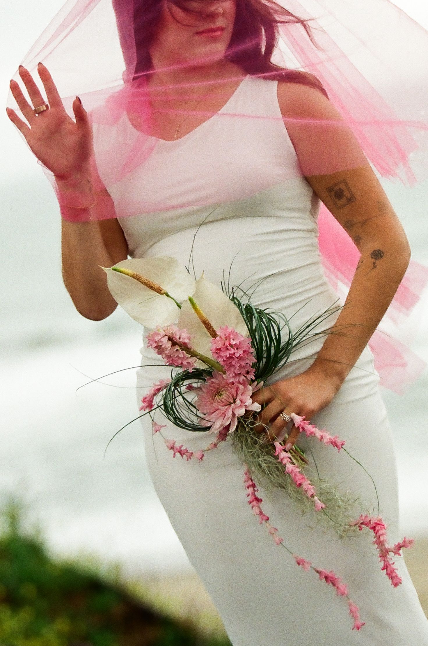Bride in white fitted dress holding pink floral bouquet with calla lily, pink veil blowing in wind.