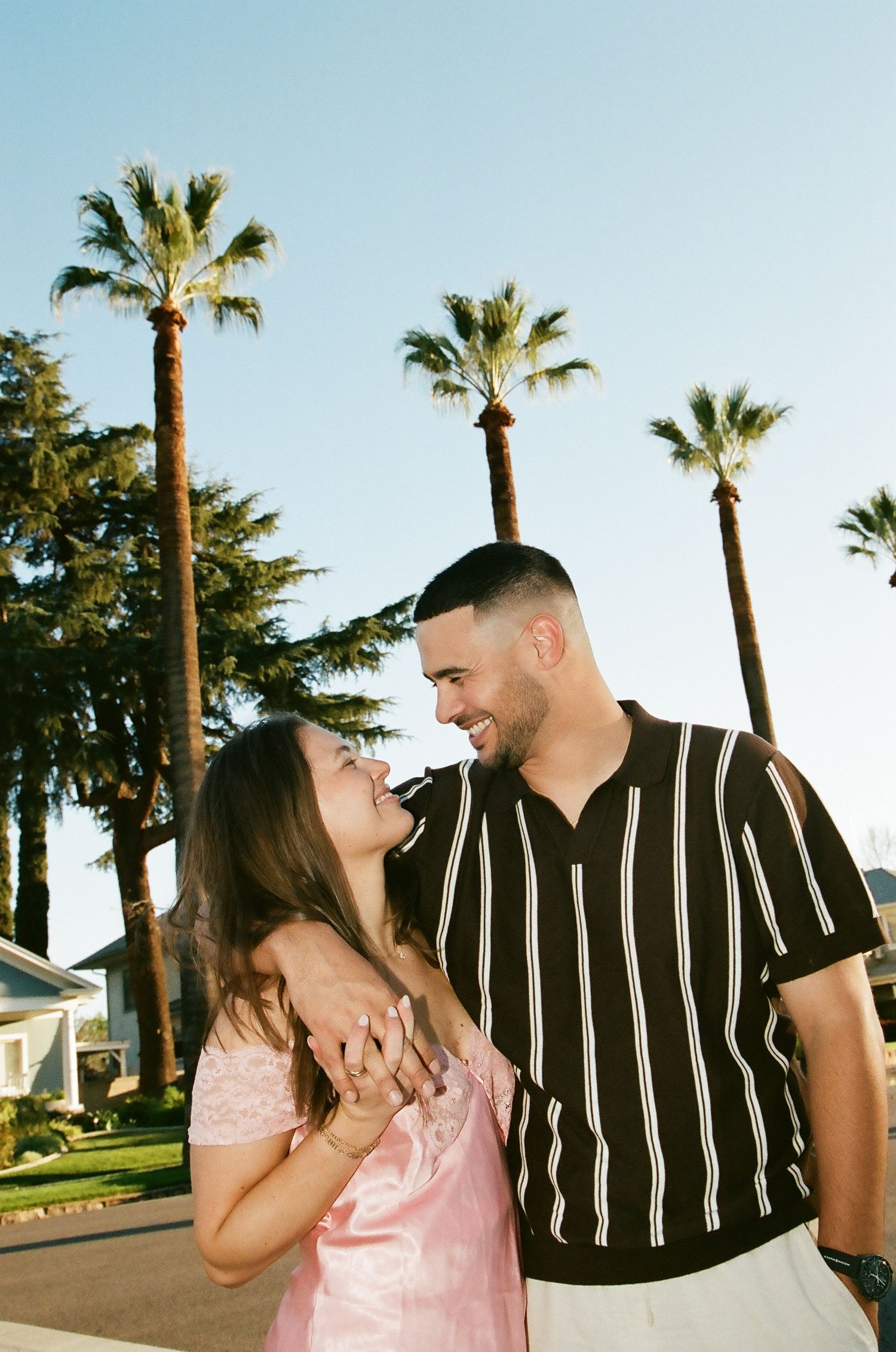 Joyful couple laughing together outdoors with tall palm trees and blue sky behind them.
