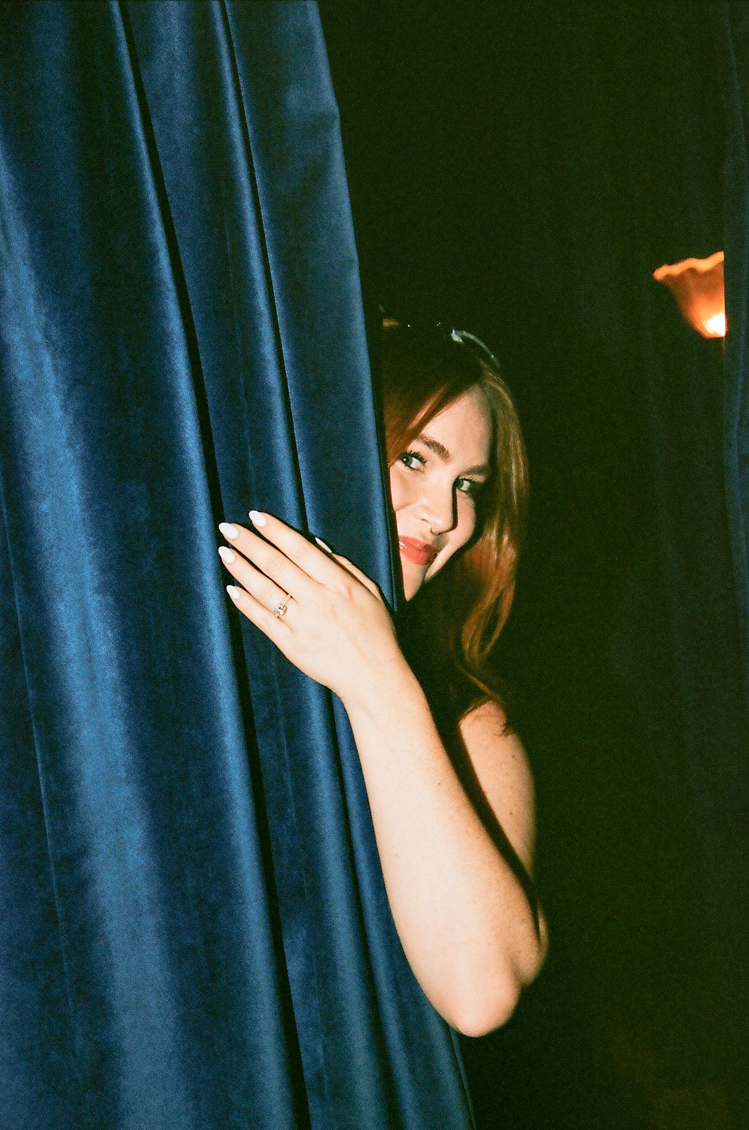 Woman peeking through blue velvet curtain, smiling and showing ring on her finger.