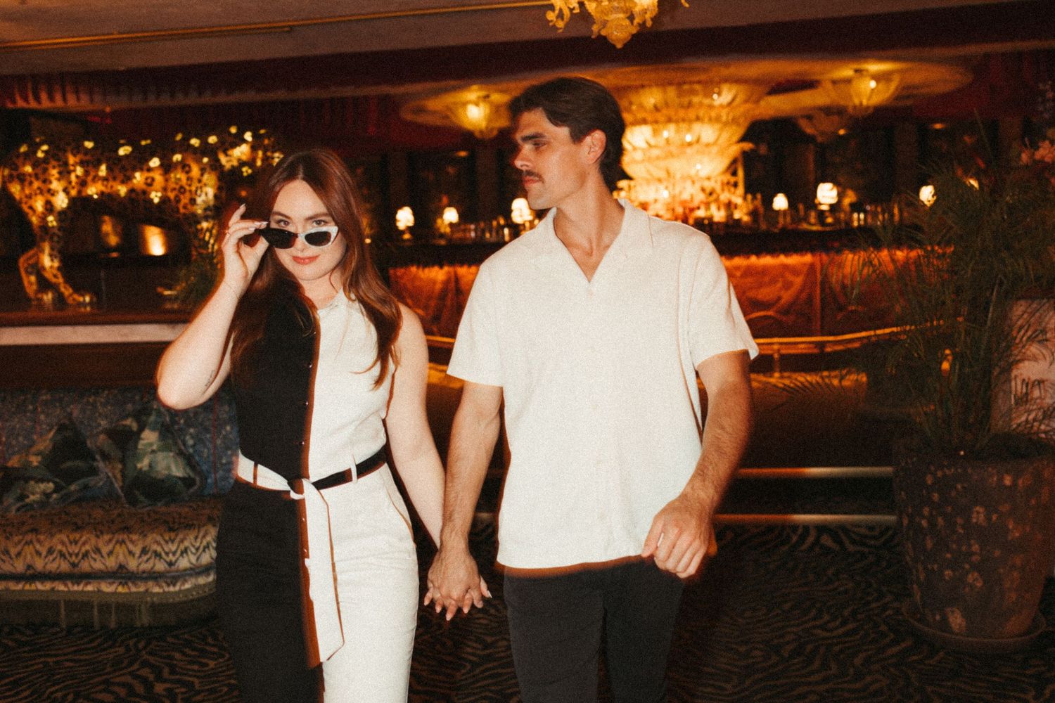 Couple holding hands in dimly lit bar, woman wearing sunglasses and white pants, man in white shirt.