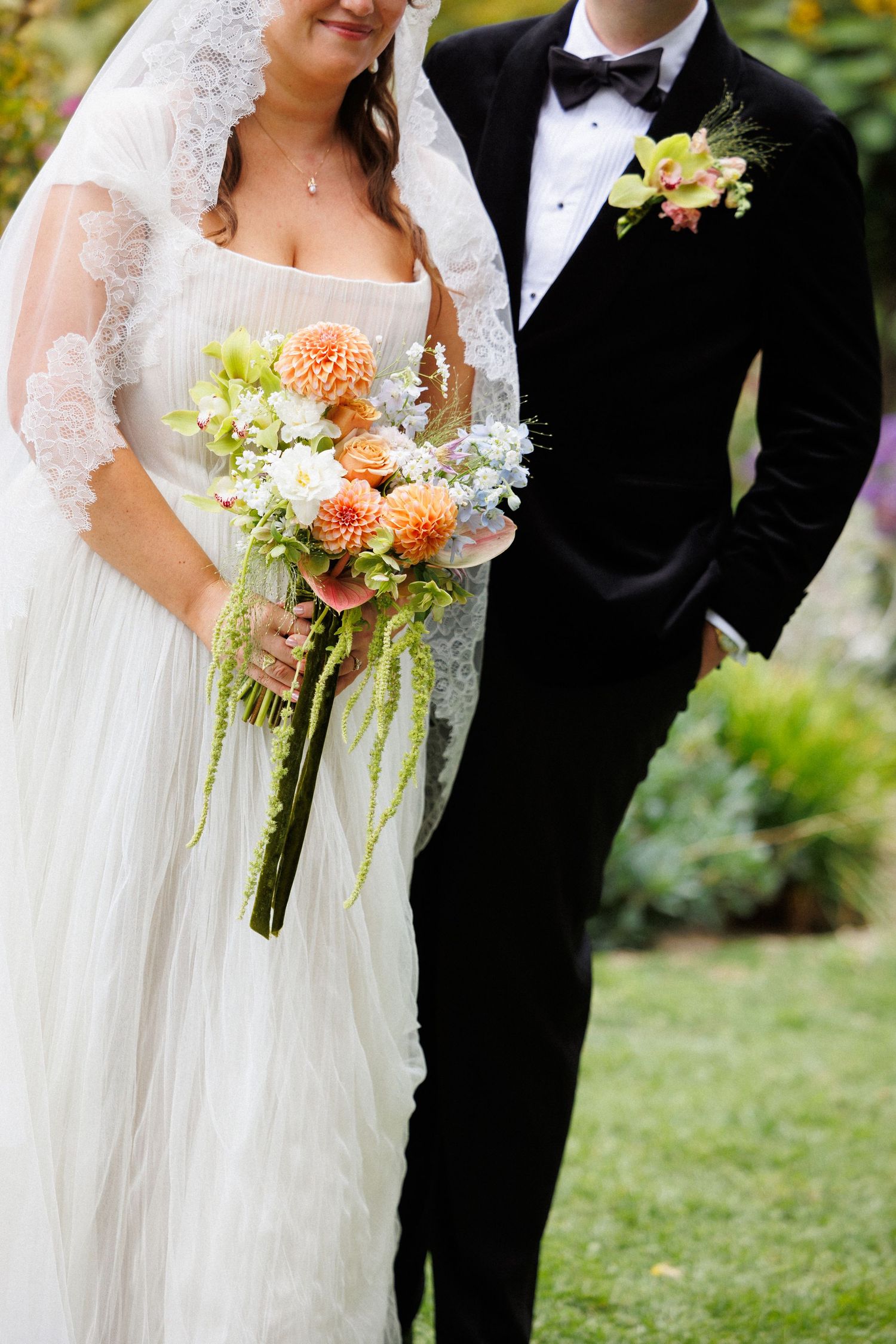 Bride and groom posing together outdoors, bride holding peach dahlia bouquet, groom in black velvet tuxedo.