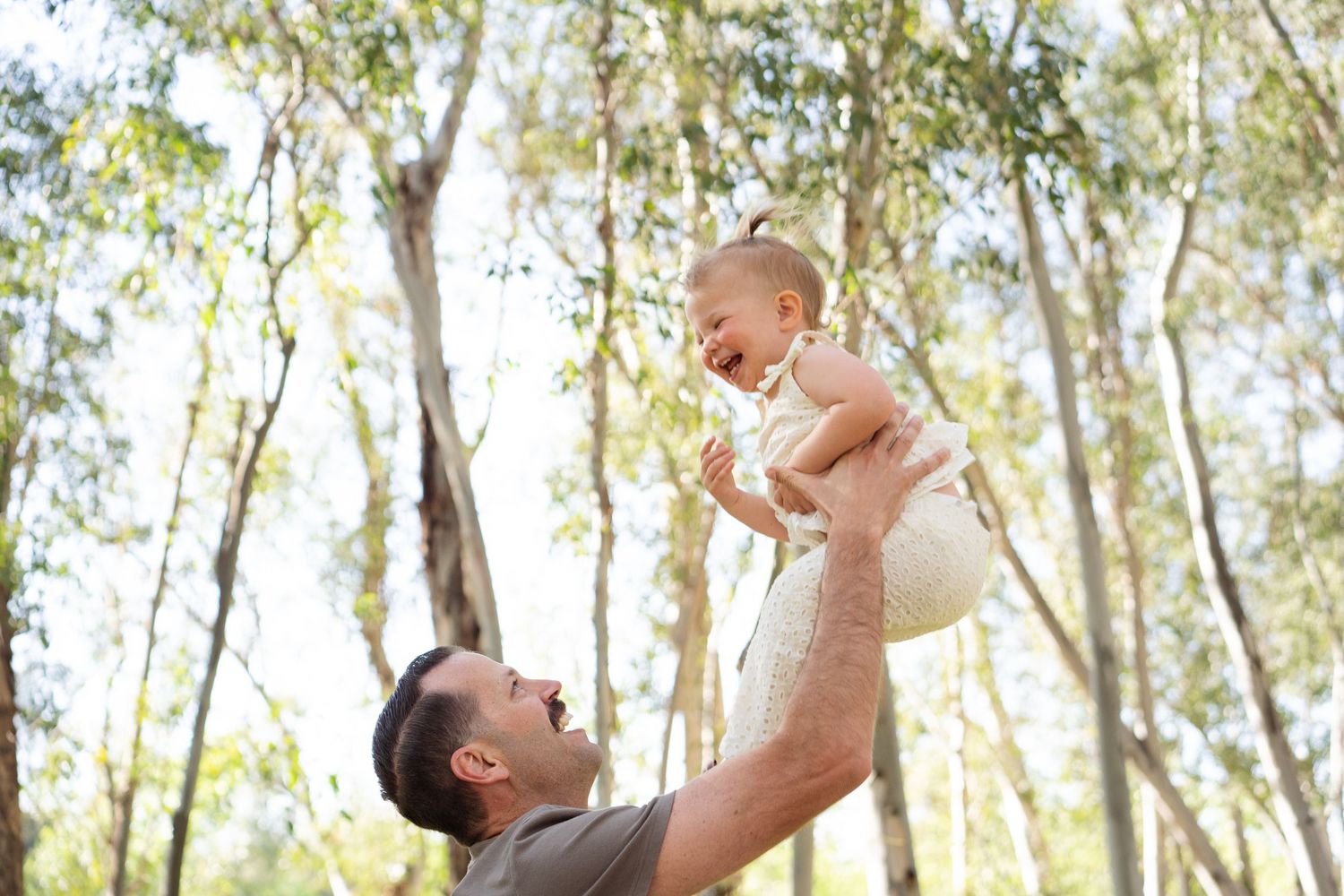 Father joyfully lifting laughing baby girl high in the air among sunlit trees outdoors.