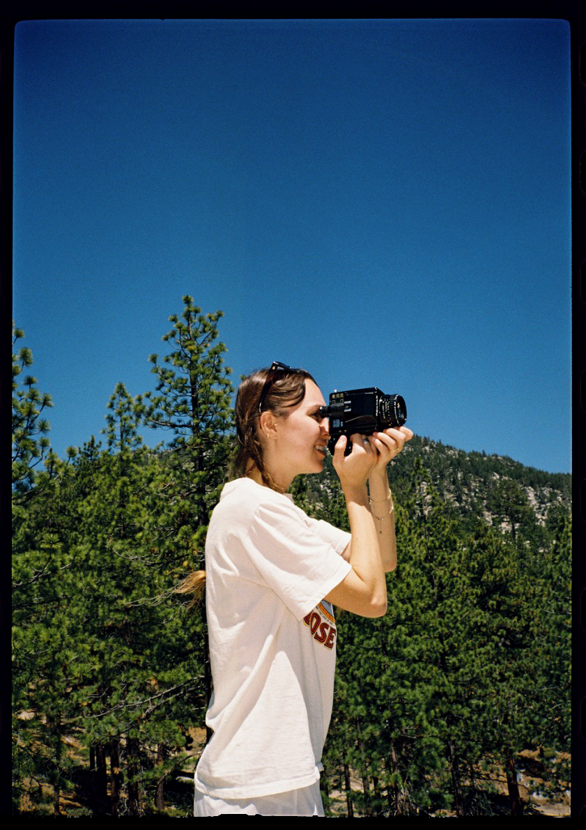 Woman in white t-shirt photographs mountain landscape with camera, pine trees and blue sky in background.