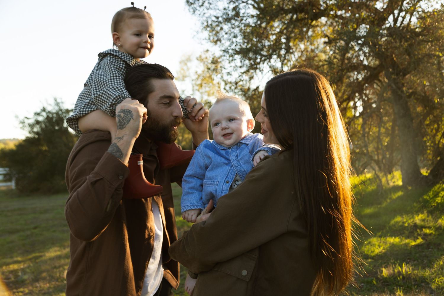 Happy family with two babies enjoying a sunny outdoor park setting, parents smiling and holding young children.