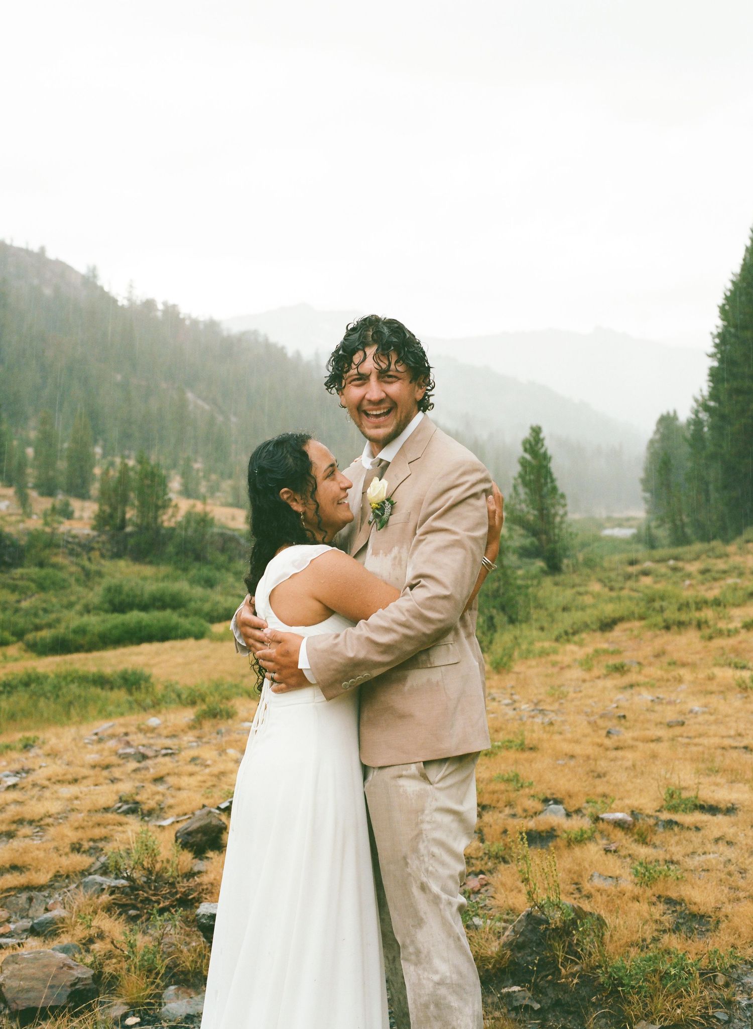 Bride and groom embracing outdoors in mountain meadow, groom laughing, pine trees and misty hills in background.