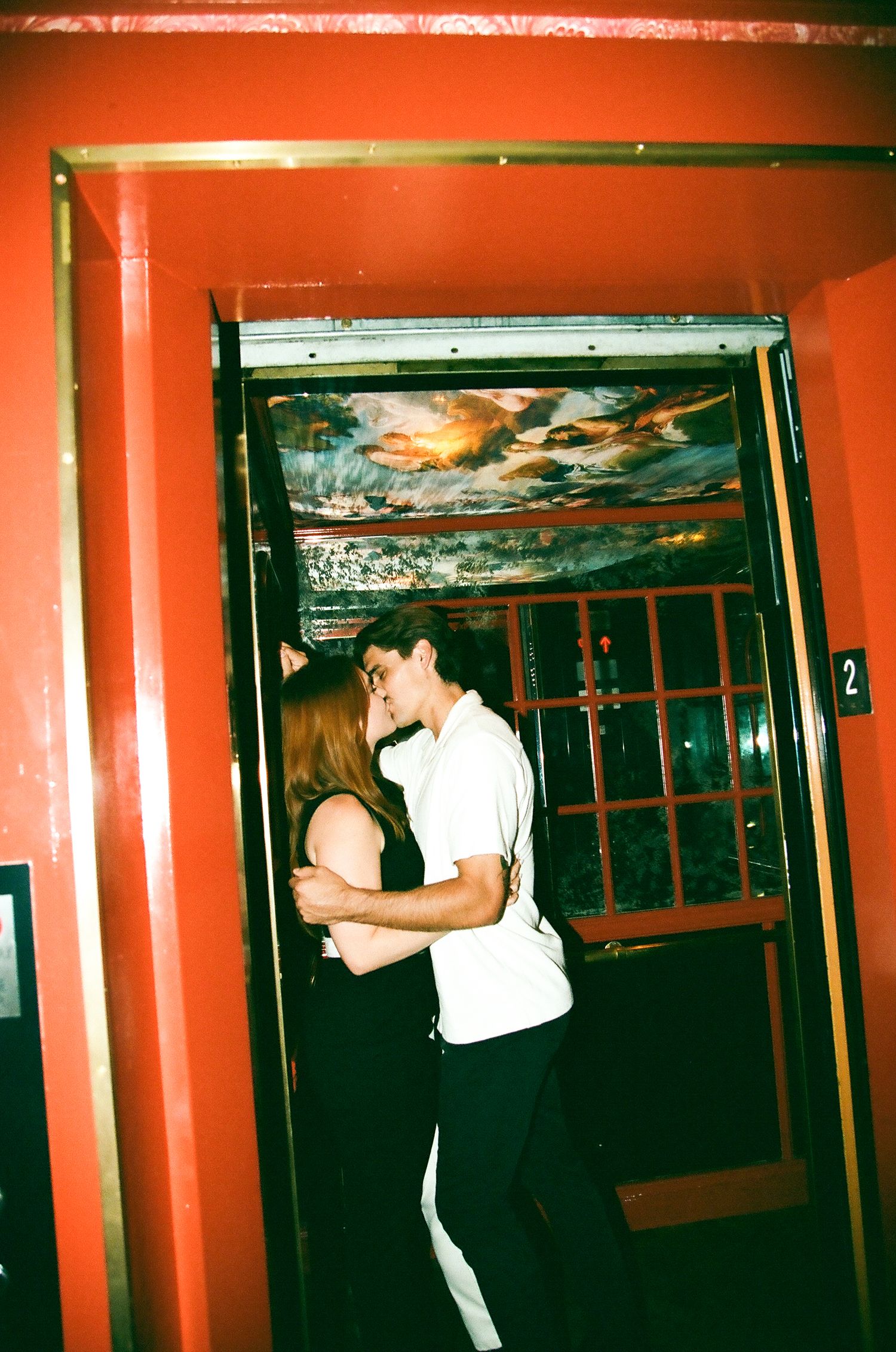 A couple embraces and kisses in a doorway with red walls and ornate ceiling painting in a vintage setting.
