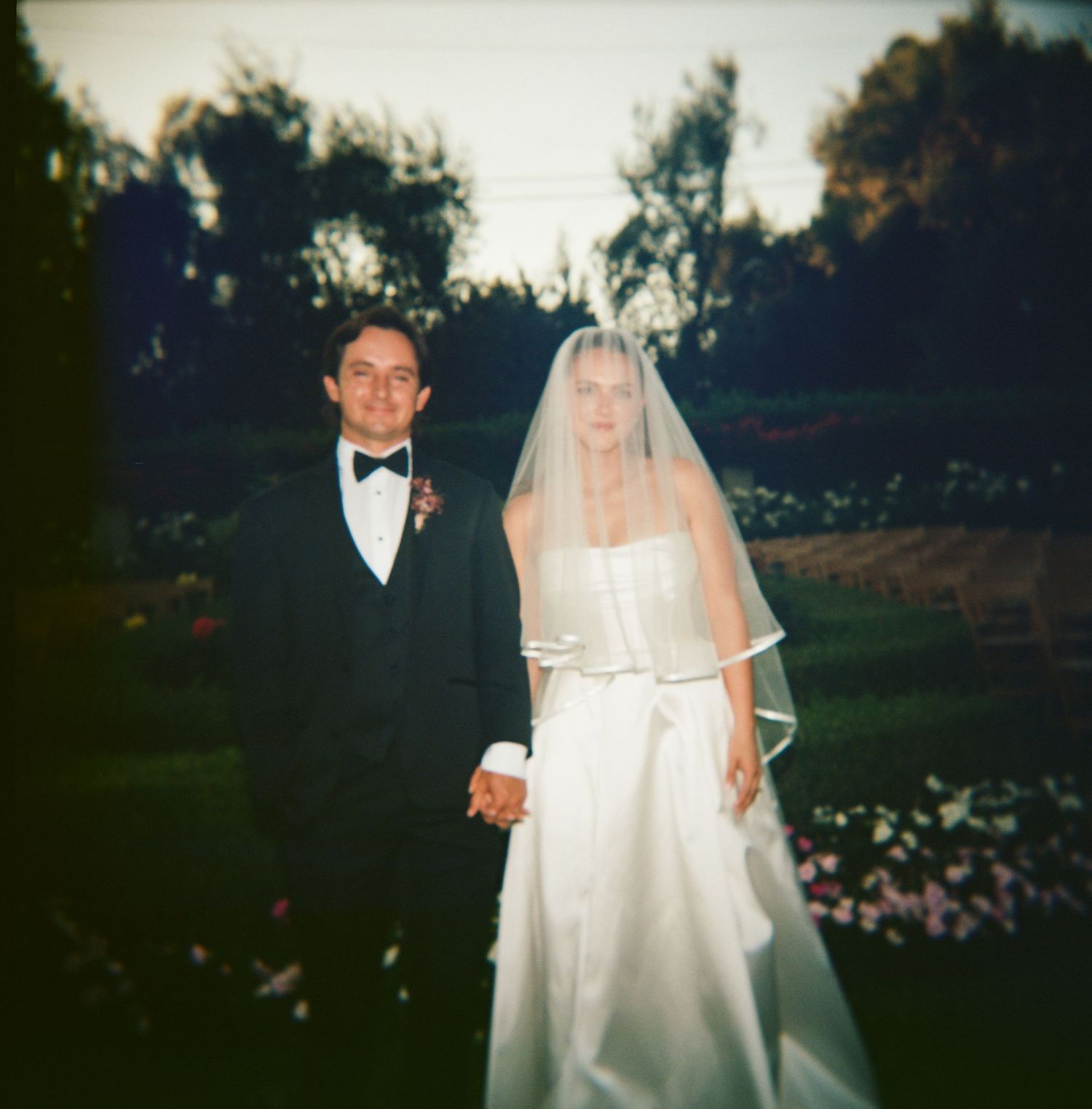 Vintage film photo of bride and groom holding hands in garden at dusk, square format.