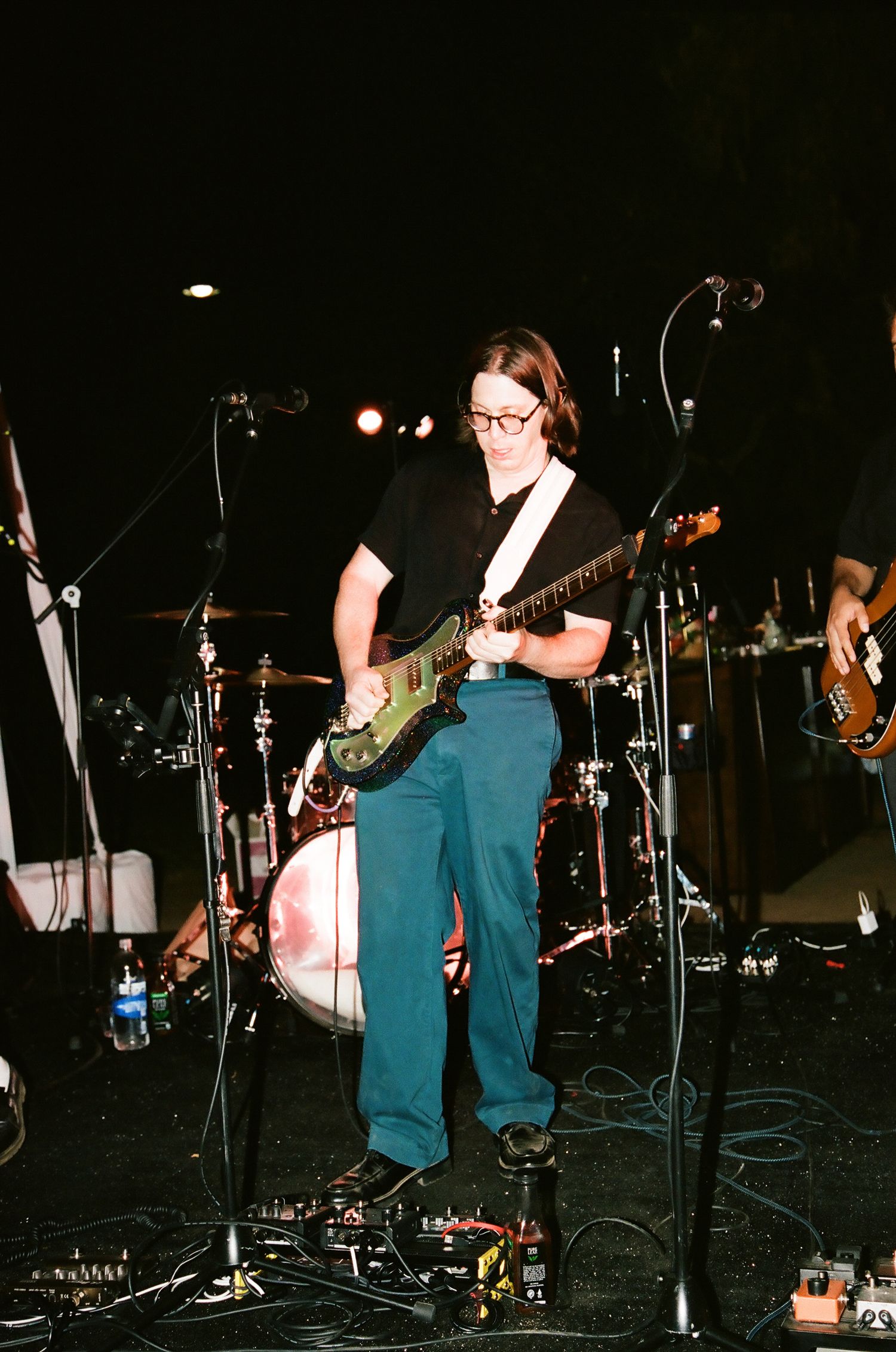 Guitarist performing on stage with teal pants, black shirt, and glasses, surrounded by microphones and guitar pedals.