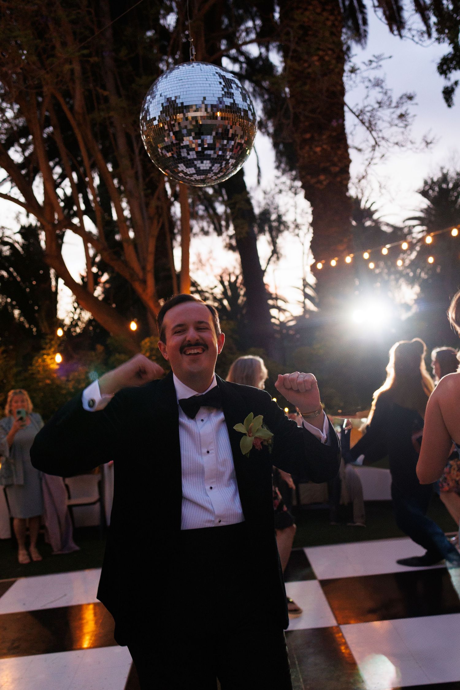 Excited groomsman in tuxedo celebrates on a checkered dance floor under a disco ball at an outdoor wedding.