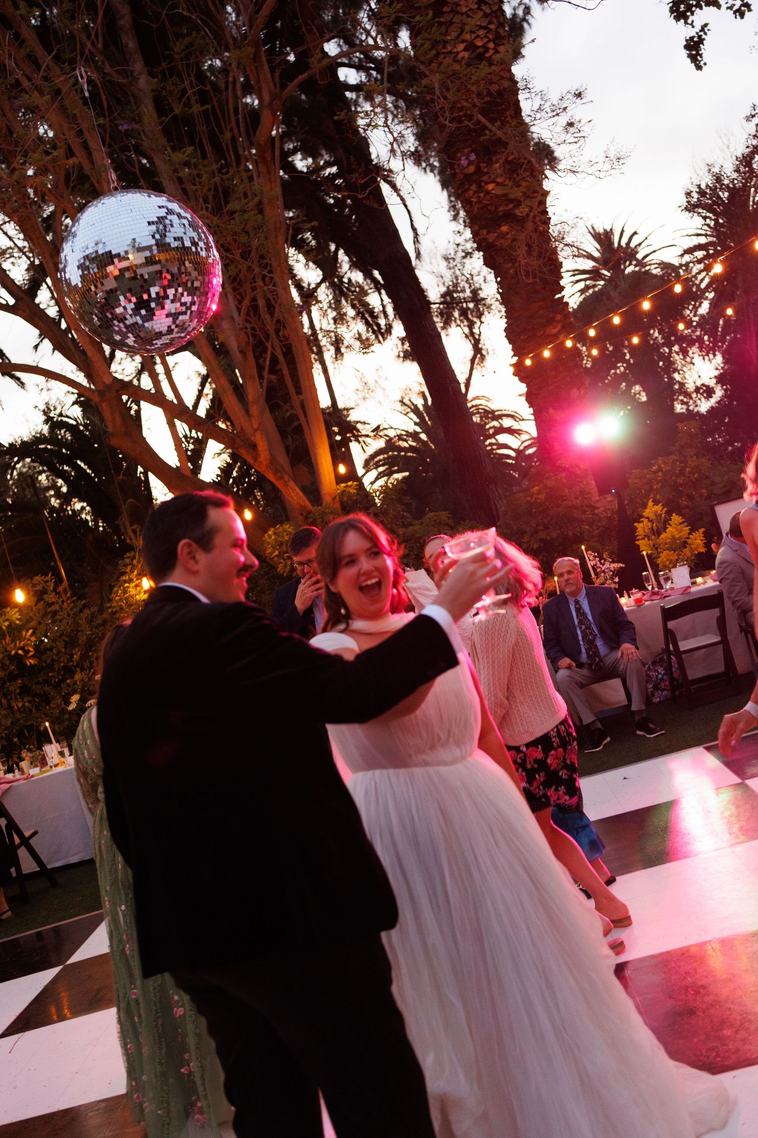 Couple dancing joyfully under a disco ball at outdoor wedding reception with pink lighting at sunset.