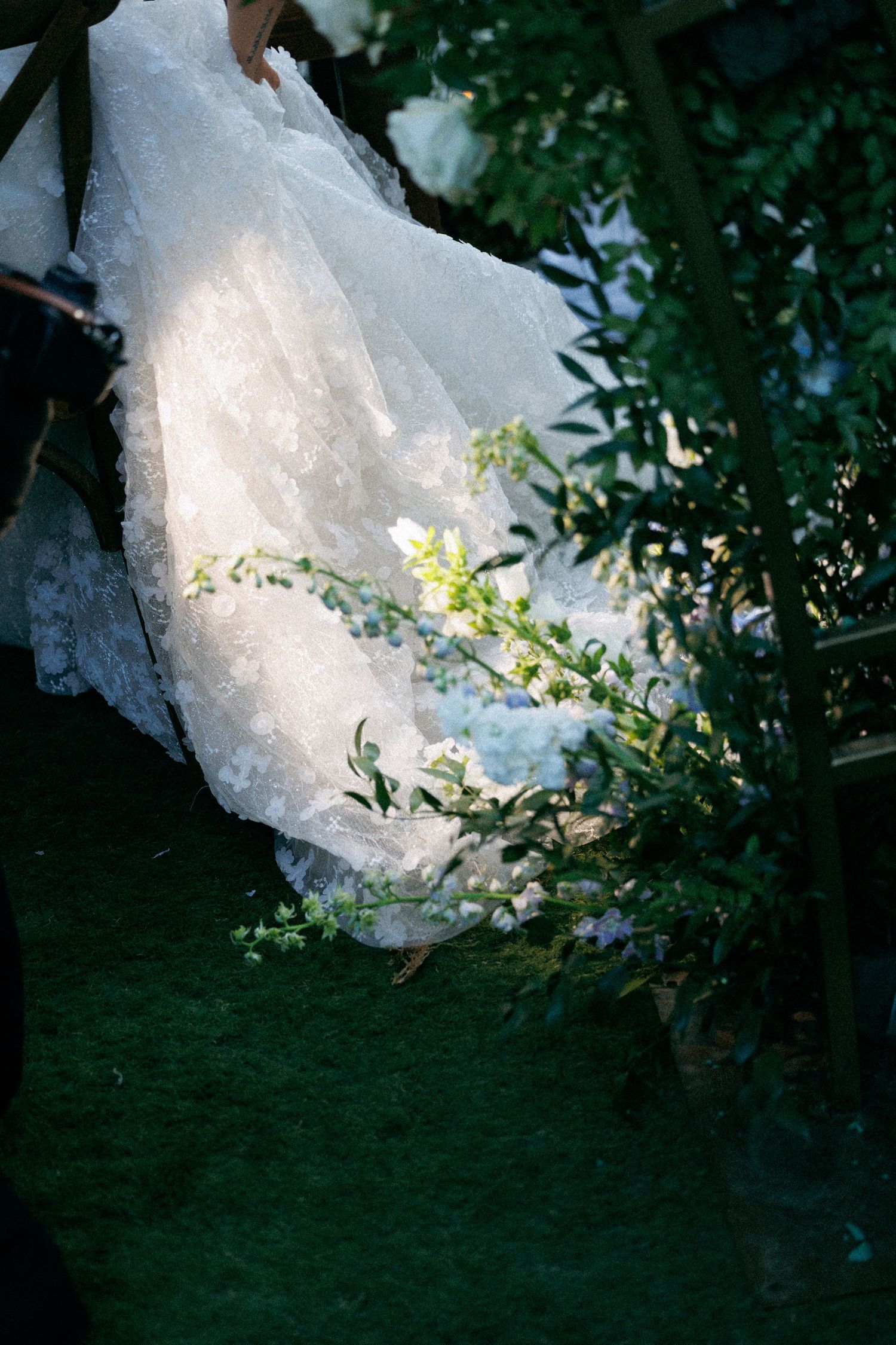 White lace wedding dress train flowing on green grass surrounded by garden foliage.