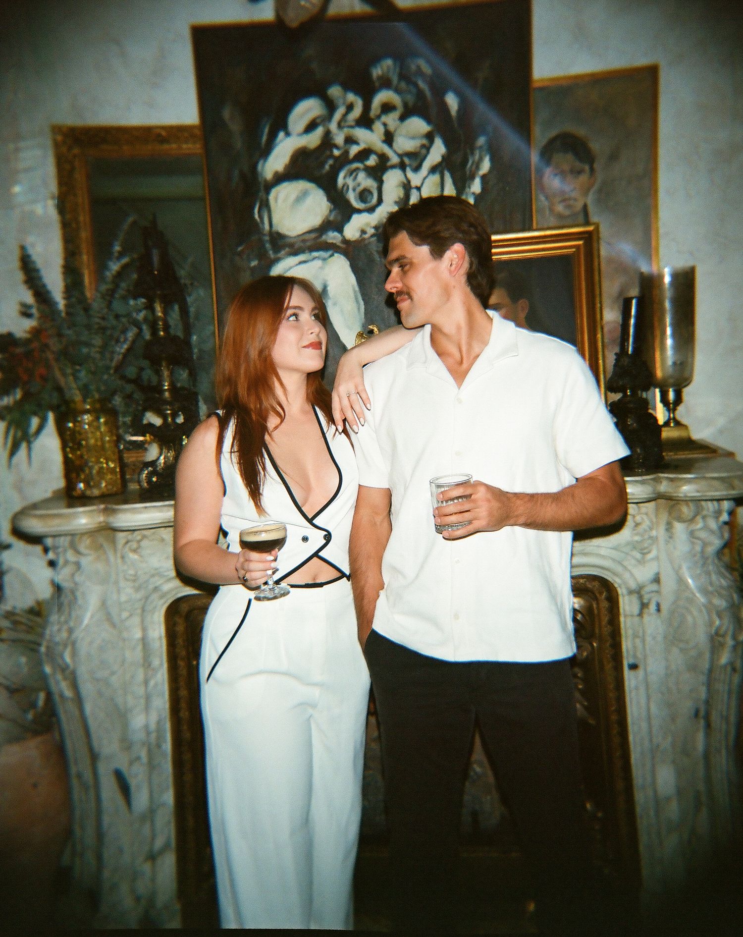 Couple holding drinks standing by ornate fireplace with artwork, woman in white jumpsuit and man in white shirt.