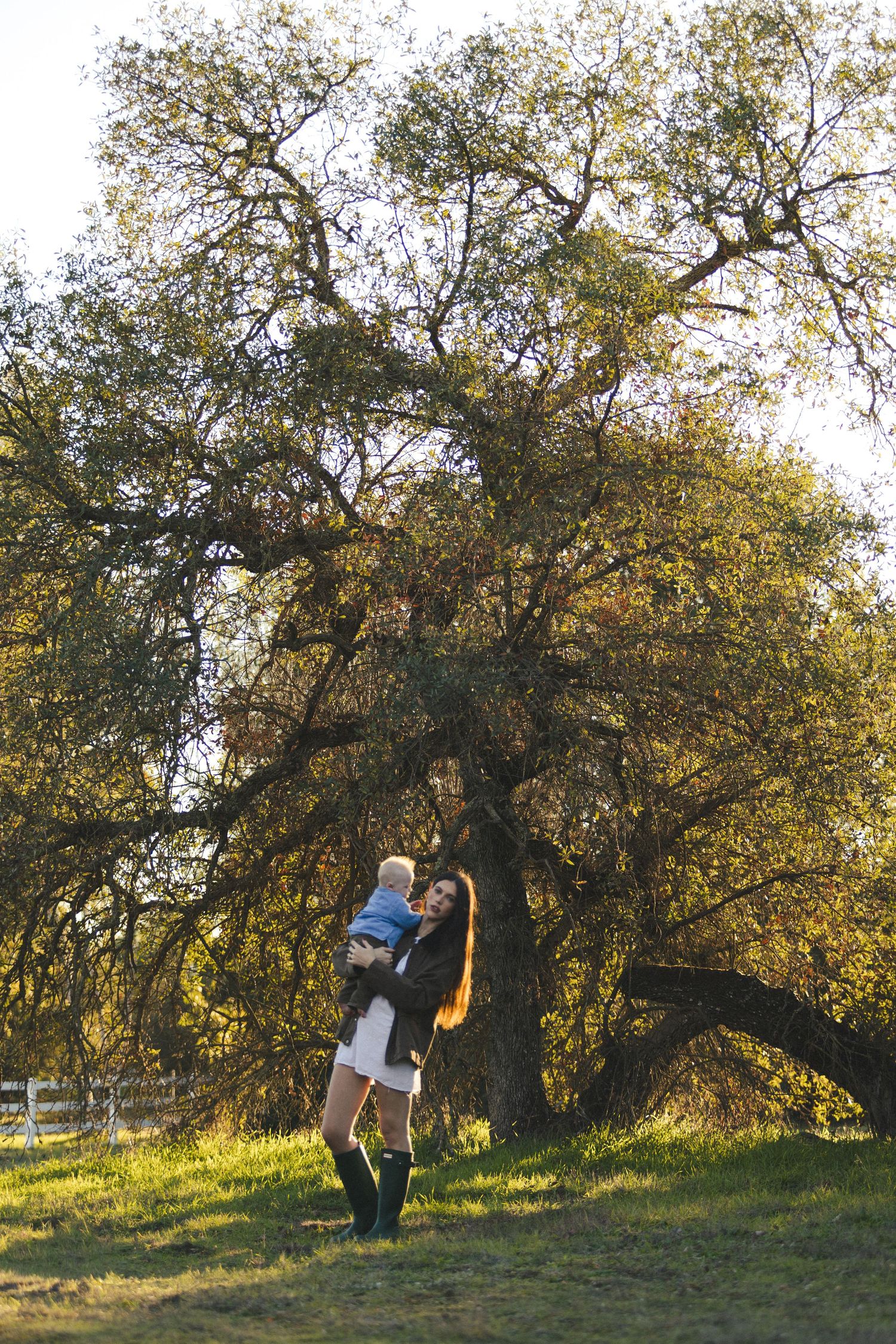 A mother lifts her baby joyfully outdoors beneath a large oak tree on a sunny day.