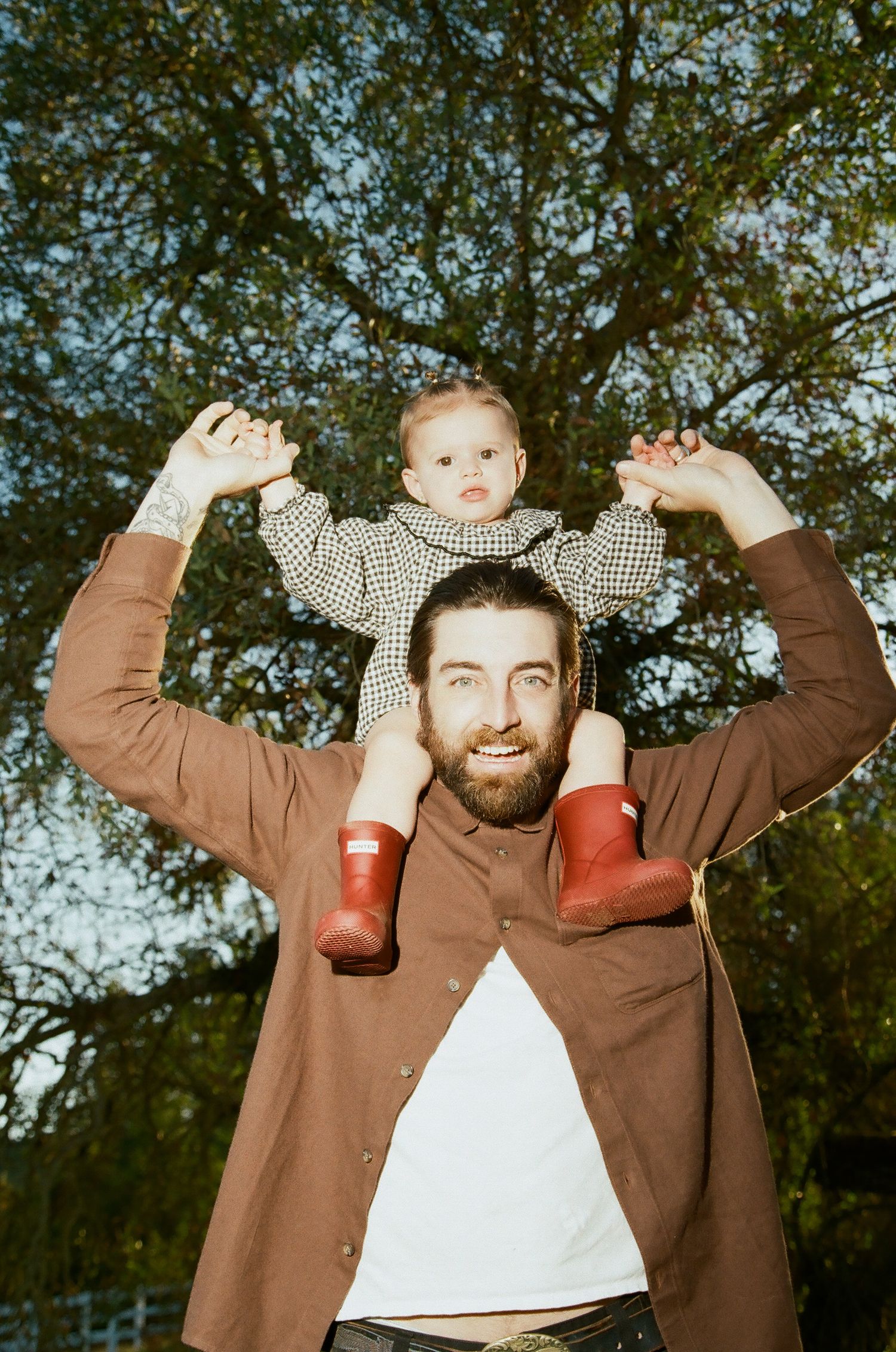 Bearded man holding toddler on shoulders outdoors, baby wearing red boots and checkered outfit, large tree background.