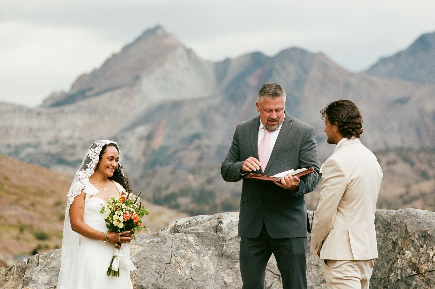 Outdoor mountain wedding ceremony with bride, groom, and officiant standing on rocks with scenic peaks in background.