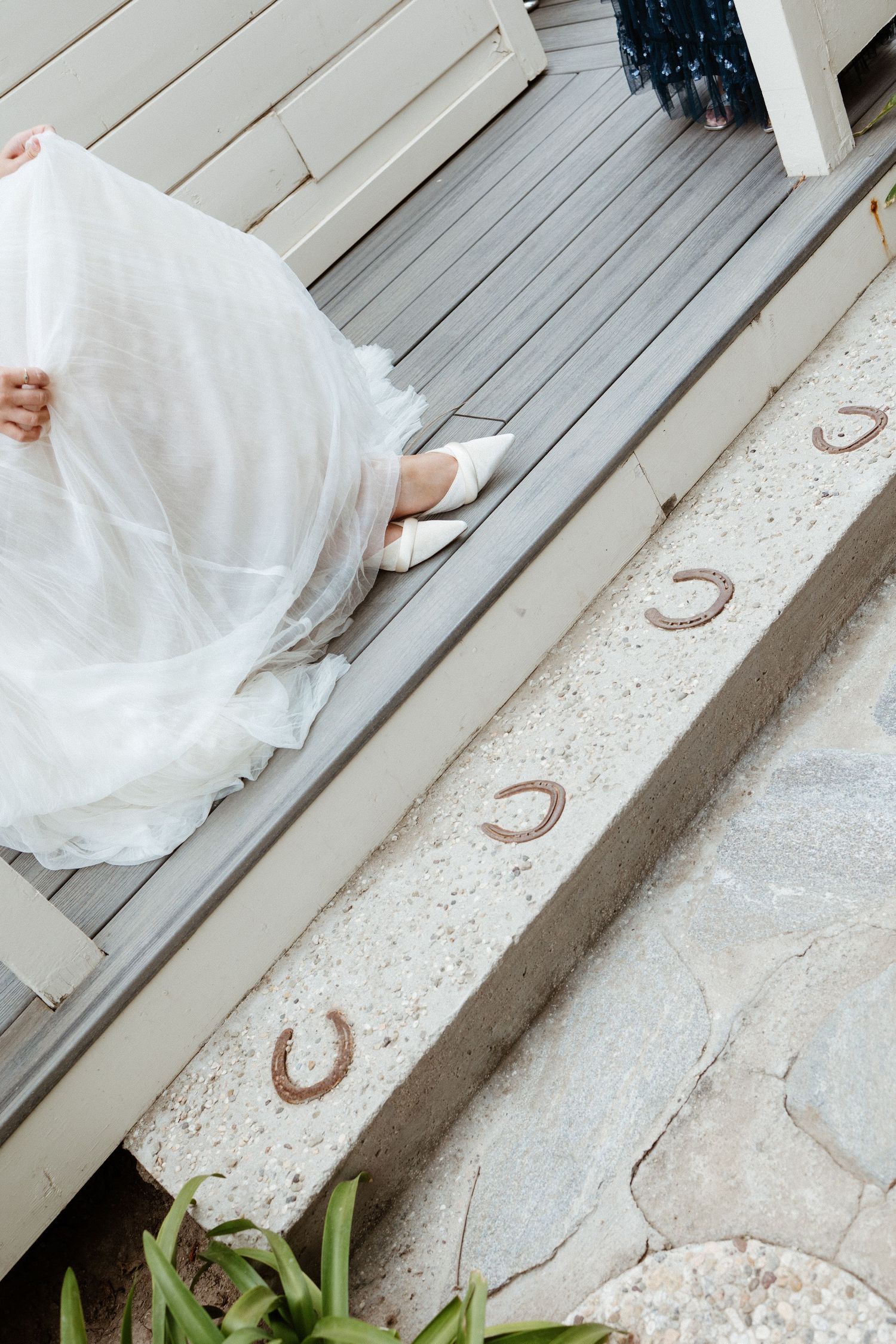 Bride in white wedding gown and heels seated on gray deck steps adorned with decorative horseshoes.