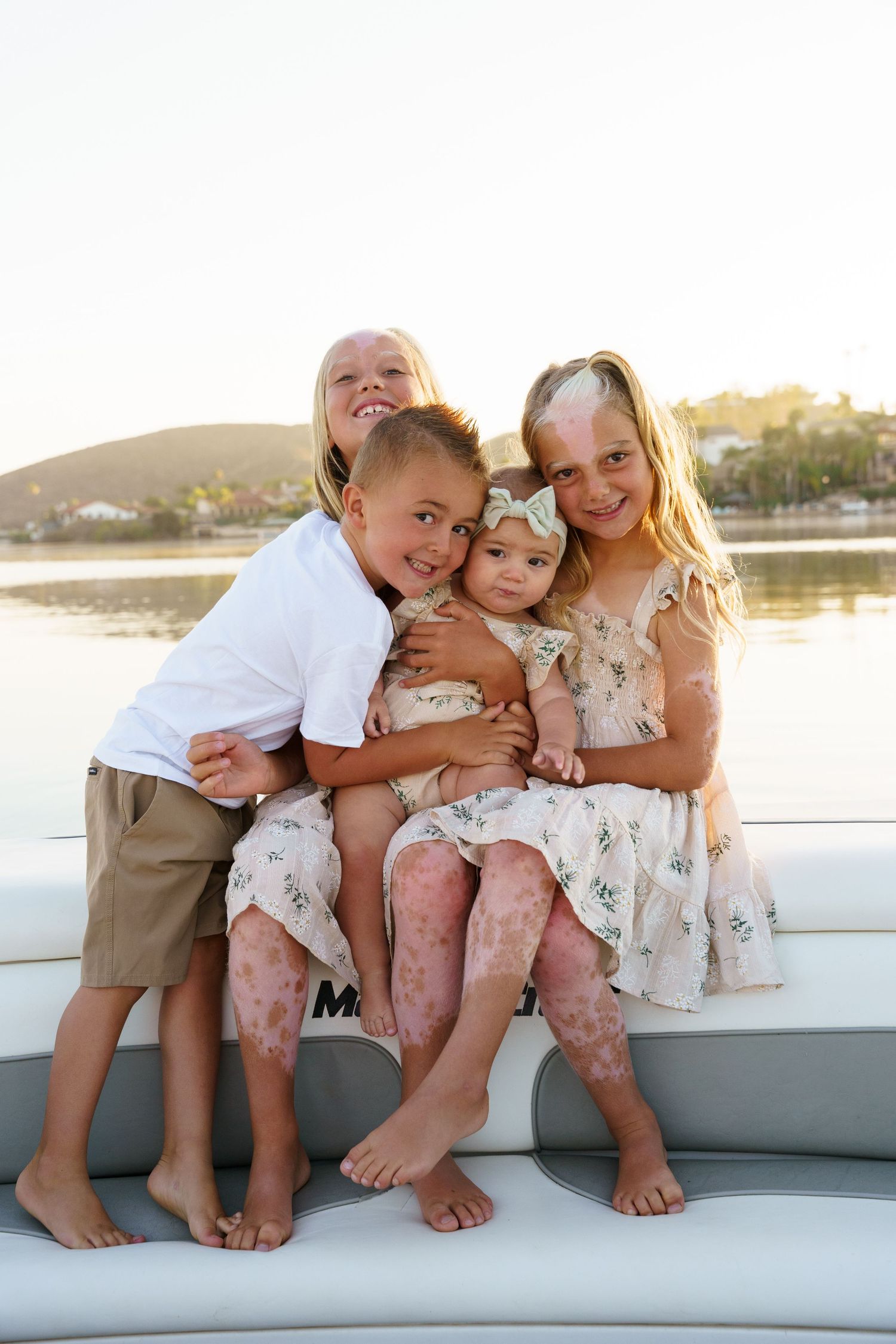 Four young siblings cuddle together on a boat at sunset, smiling for a portrait by the water.