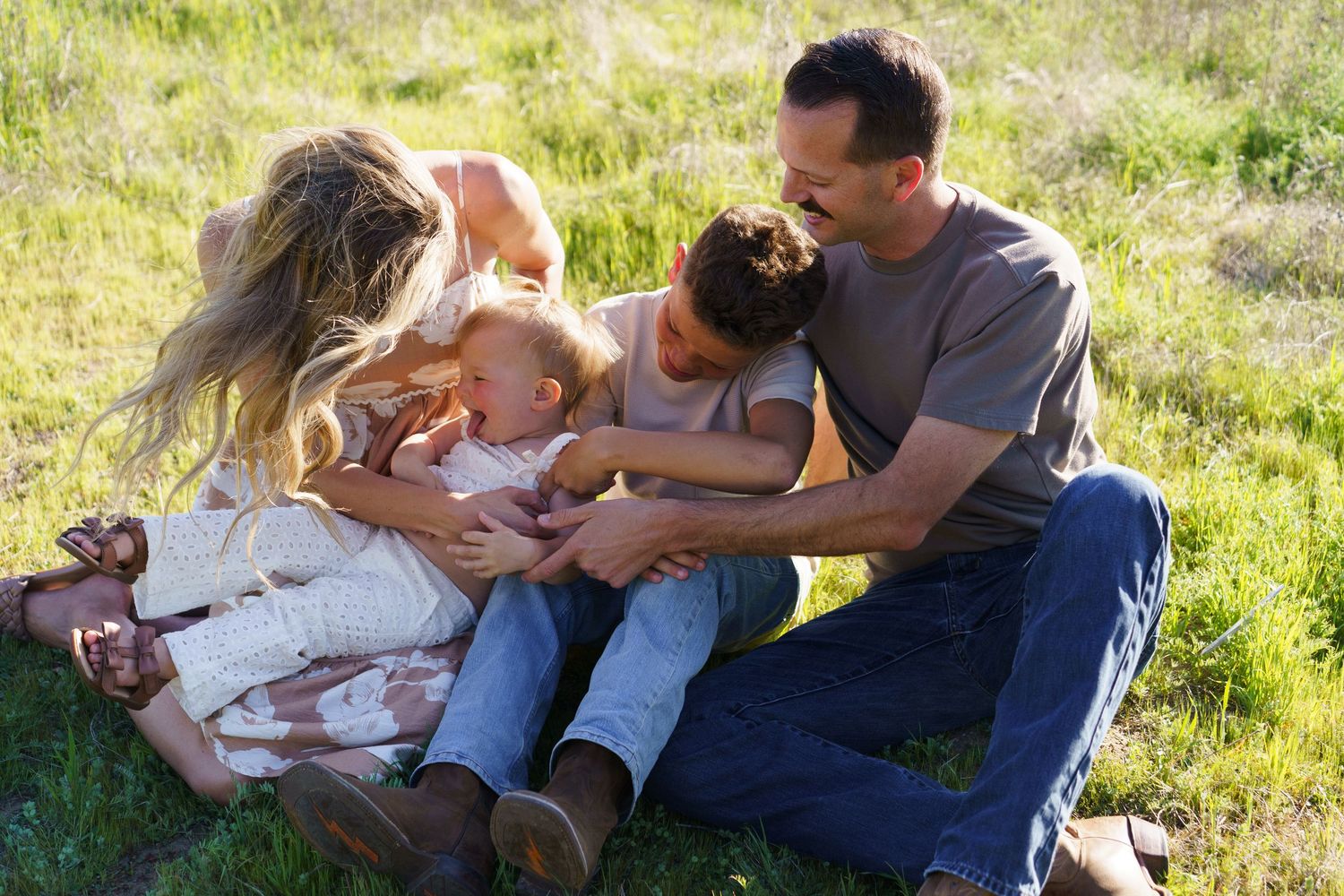 Loving family of four sitting in green grass, parents cuddling two young children outdoors.