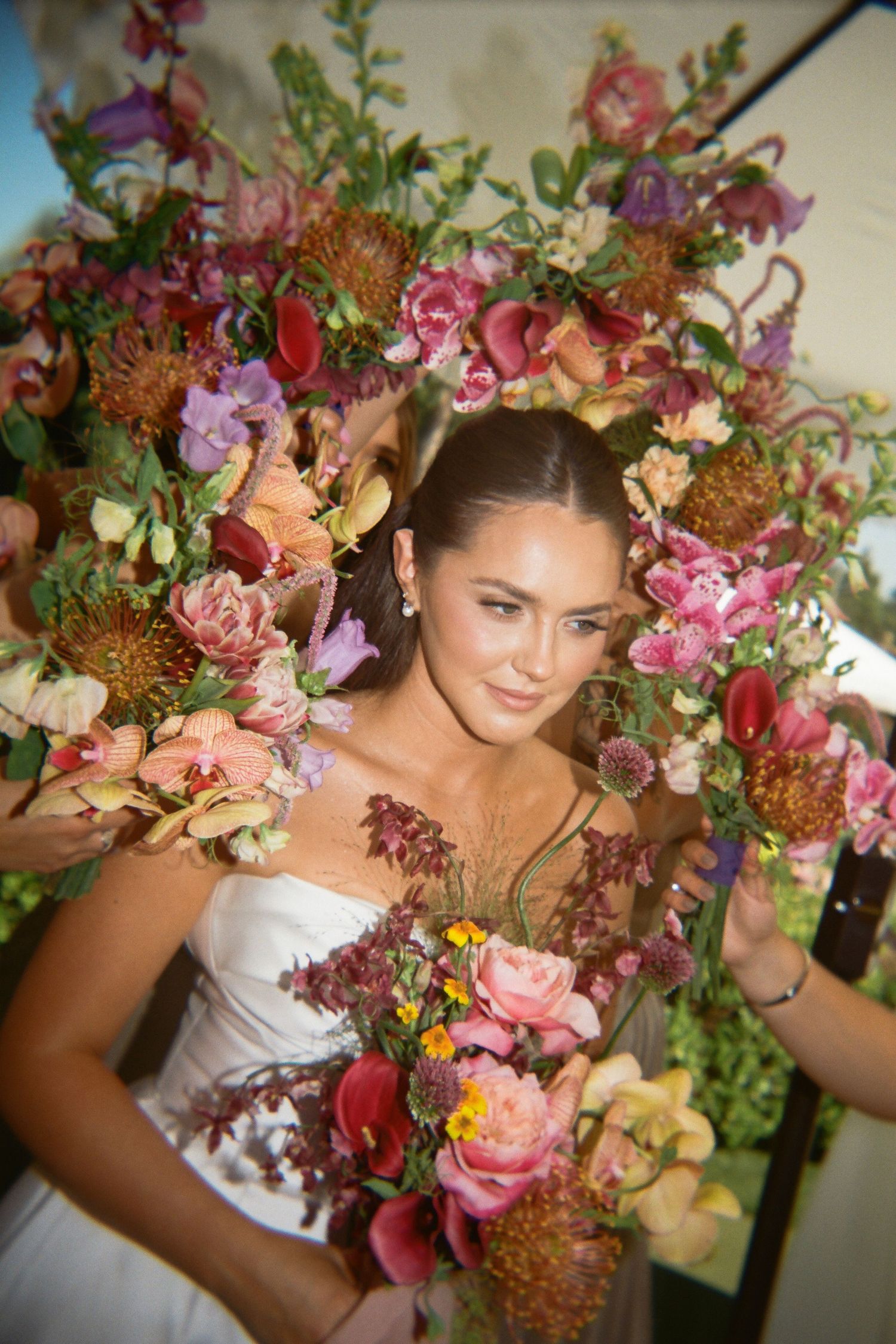 Bride surrounded by a lush floral wreath of pink, red, and orange blooms, holding a matching bouquet.