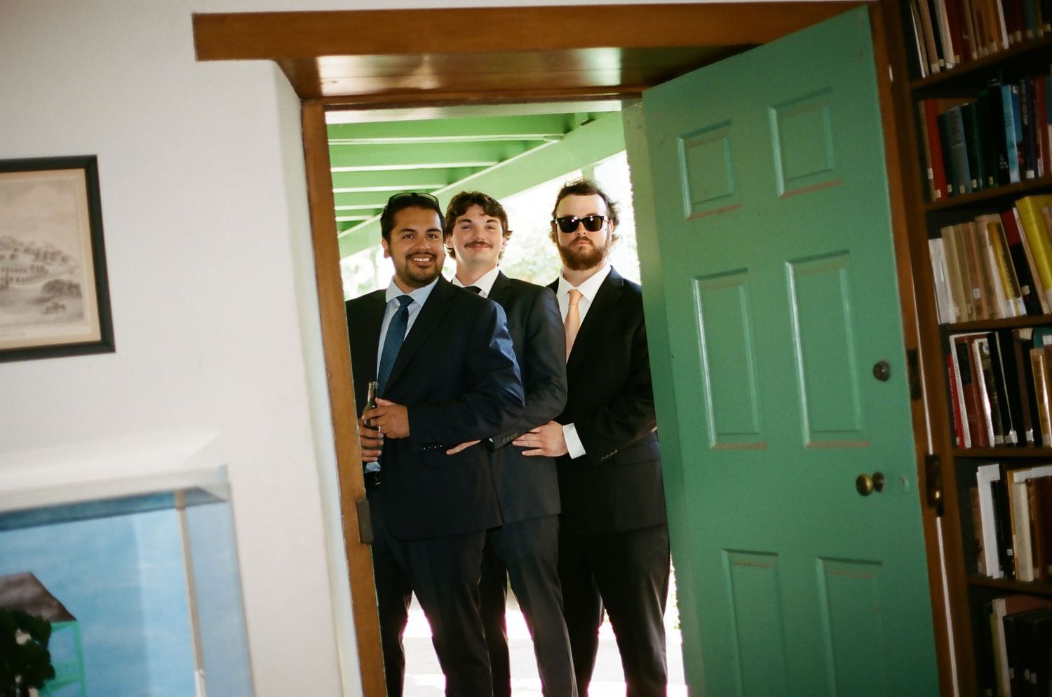 Three men in suits and ties stand in a doorway next to a bookshelf at a formal event.
