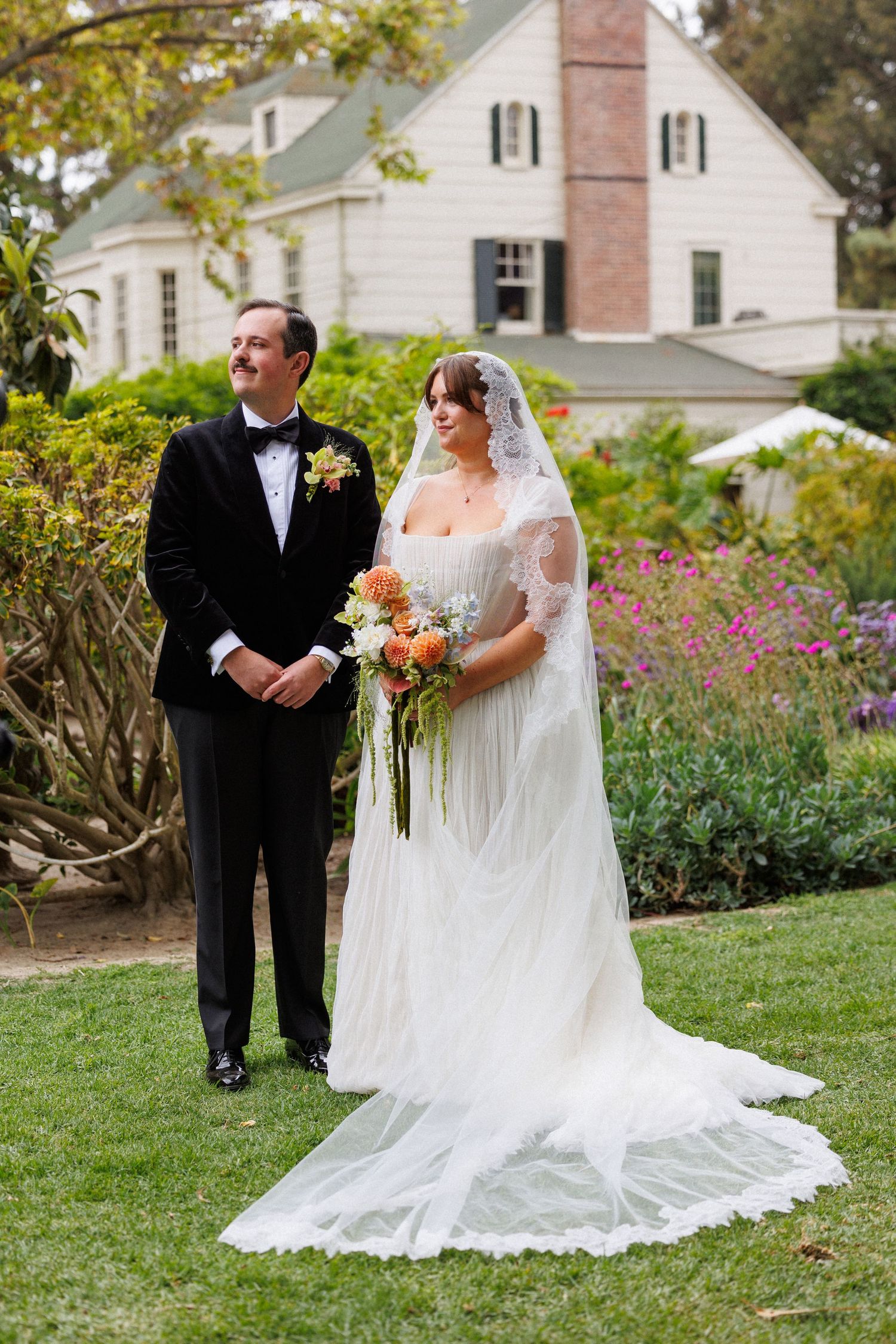 Bride in lace veil with bouquet and groom in tuxedo pose on lawn before charming white farmhouse.