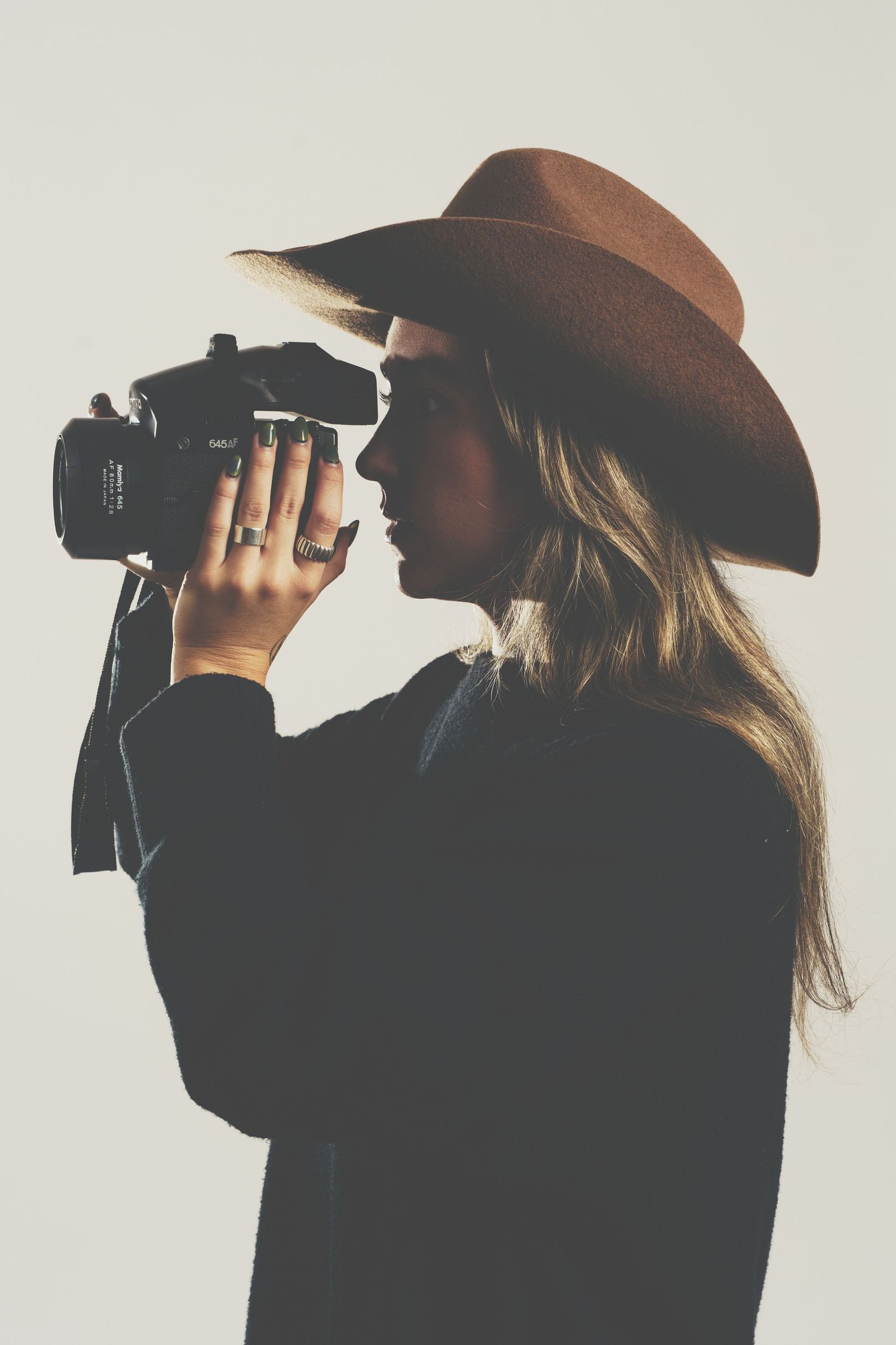 Stylish woman in wide-brim brown hat photographing with a film camera against a light background.