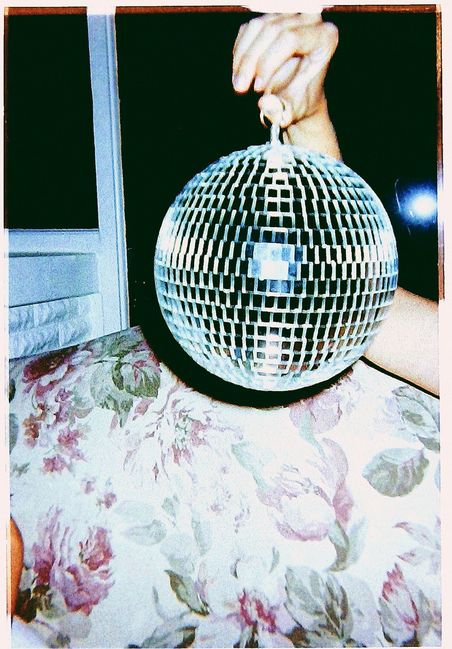A hand holds a shiny disco ball above a floral patterned surface indoors.