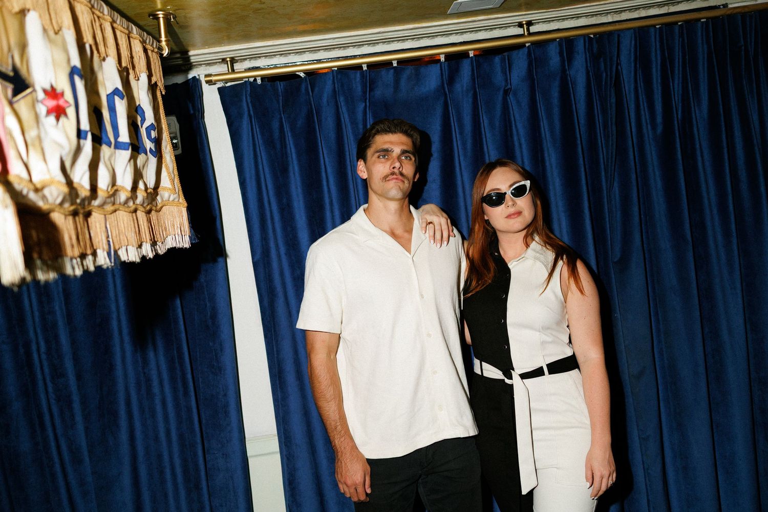 Couple posing together in front of blue velvet curtain backdrop, woman in black and white outfit.