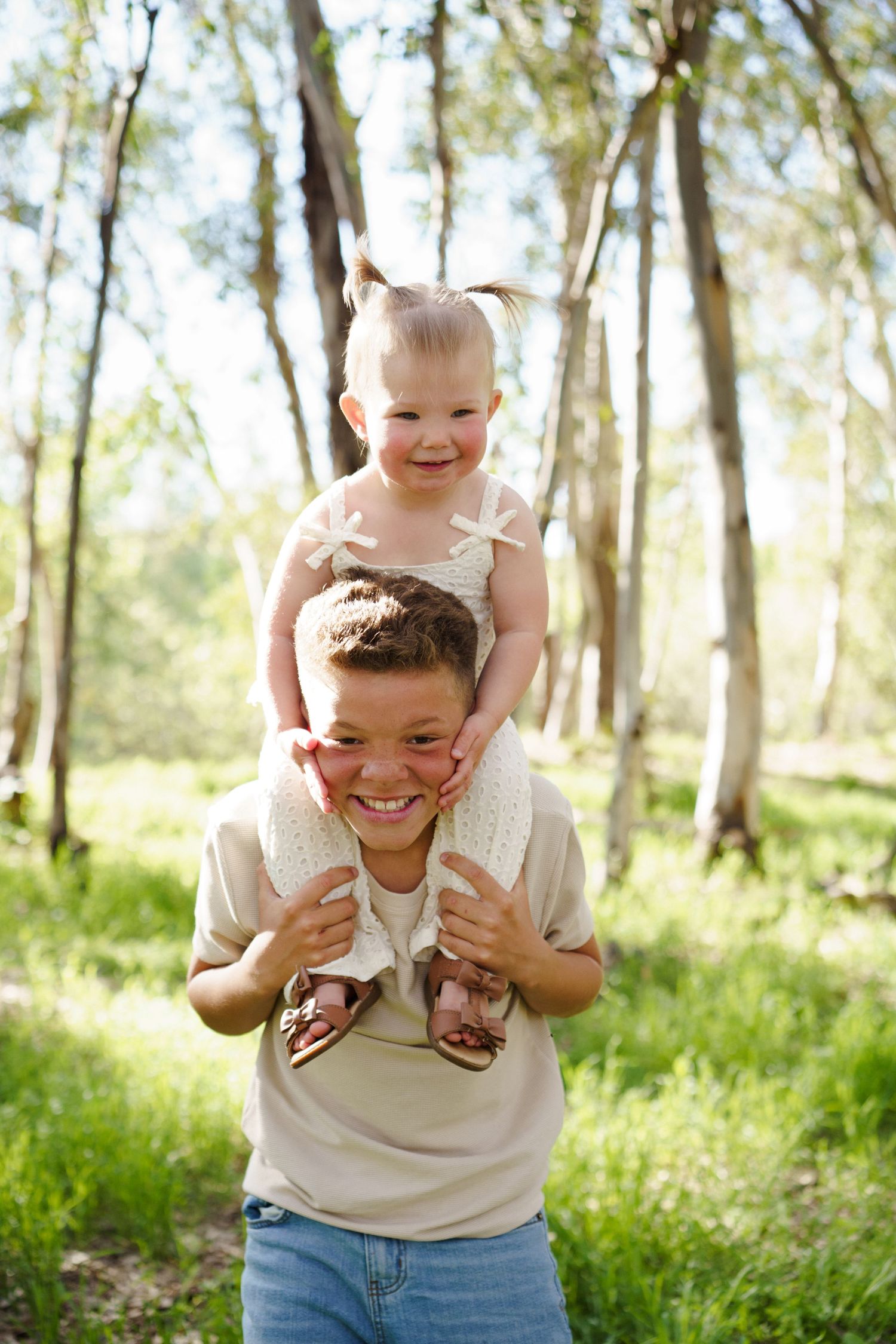 Smiling boy carrying giggling toddler sister on shoulders in bright sunlit forest setting.