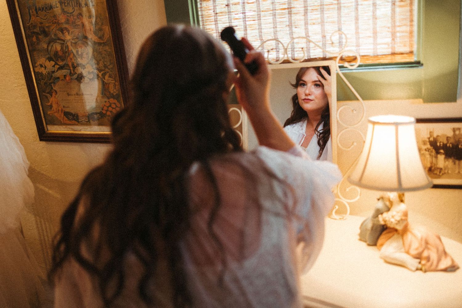 Bride in white robe styles her hair at a warmly lit vanity mirror while preparing for her wedding.