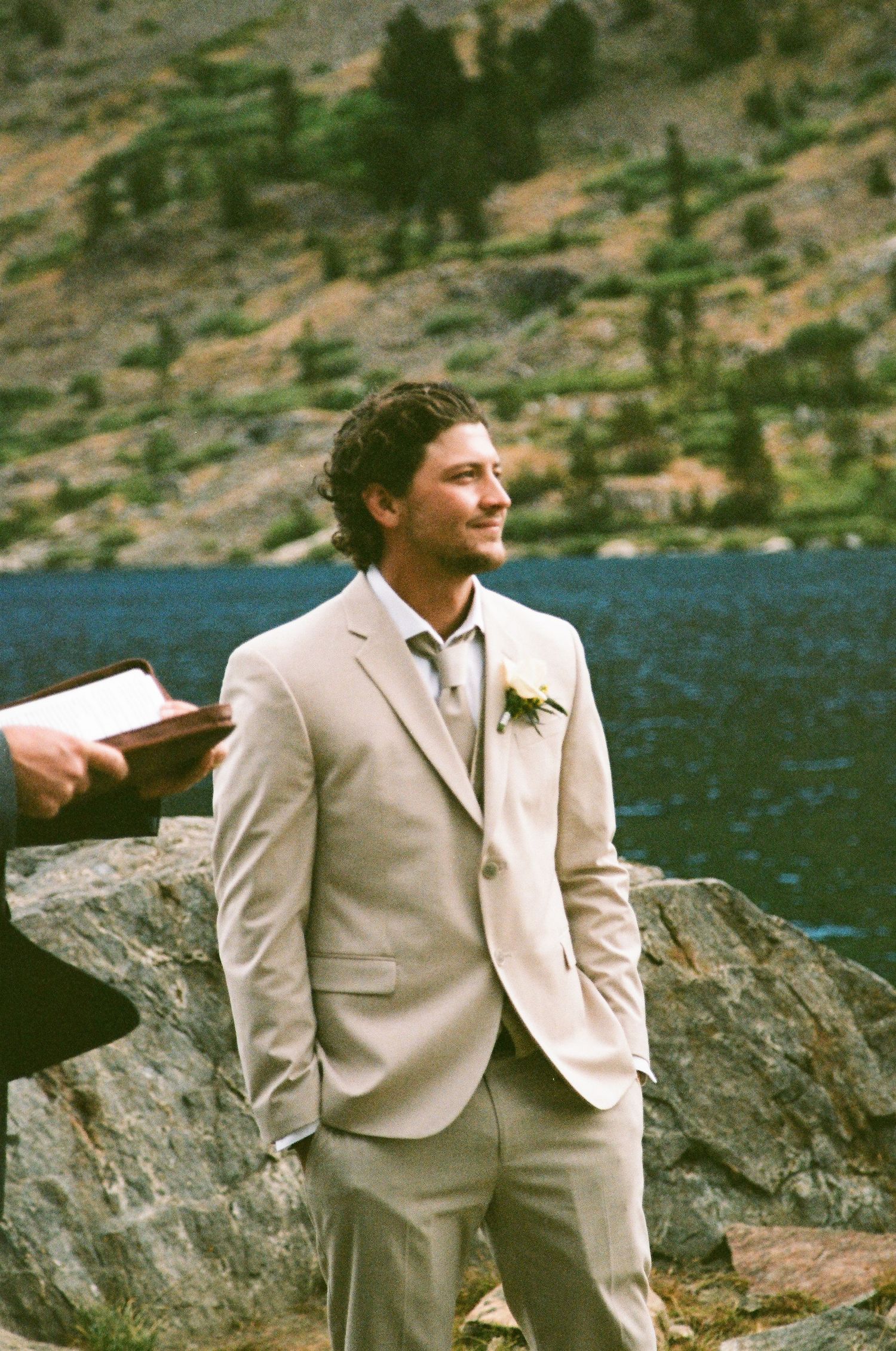 Groom in beige suit with boutonniere smiling outdoors by a mountain lake during a wedding ceremony.