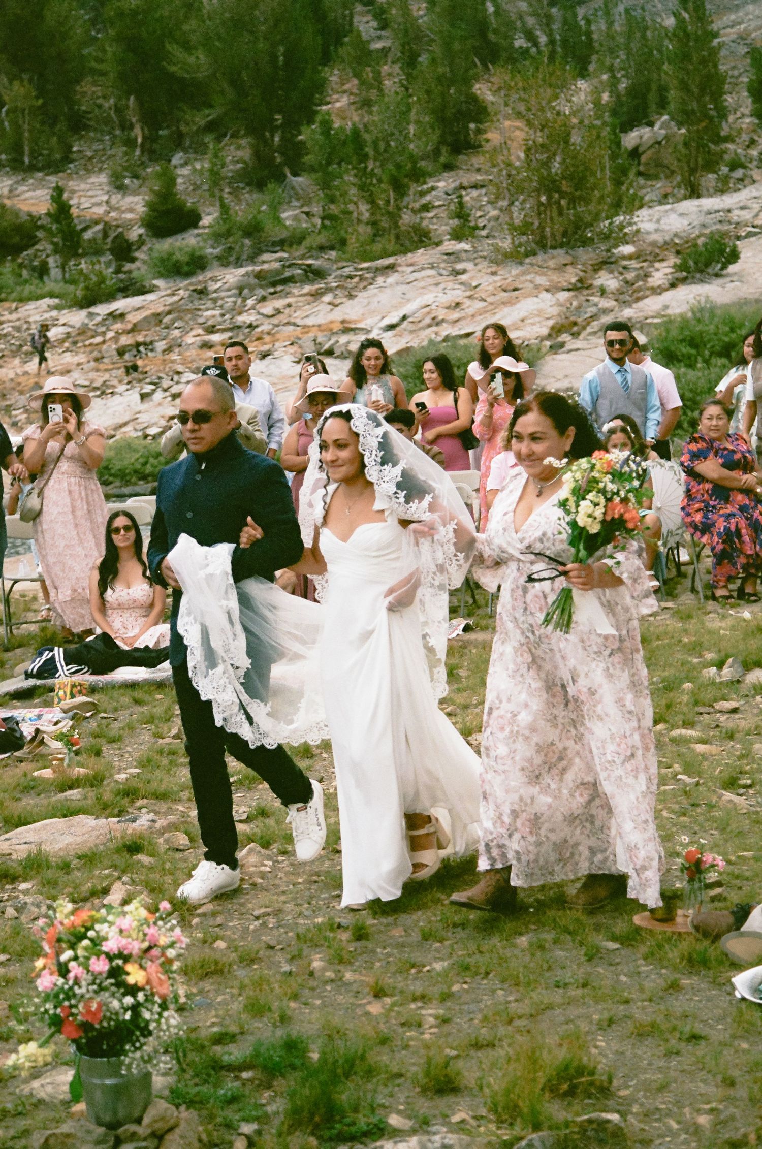 Bride in white gown and veil walks down outdoor aisle with man in black and woman in floral dress.