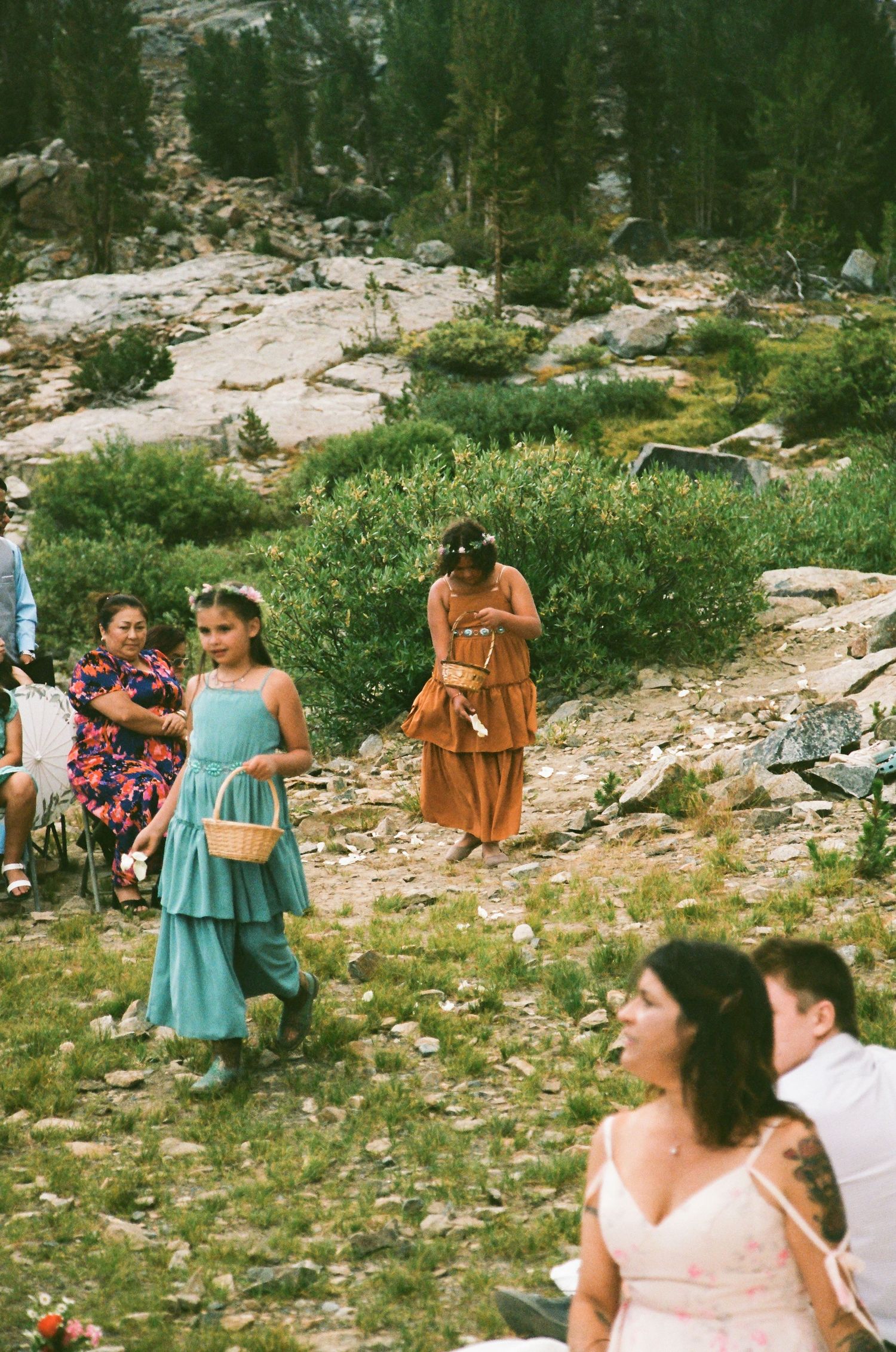 Flower girl in teal dress carries basket at outdoor mountain wedding ceremony with guests seated on rocky terrain.