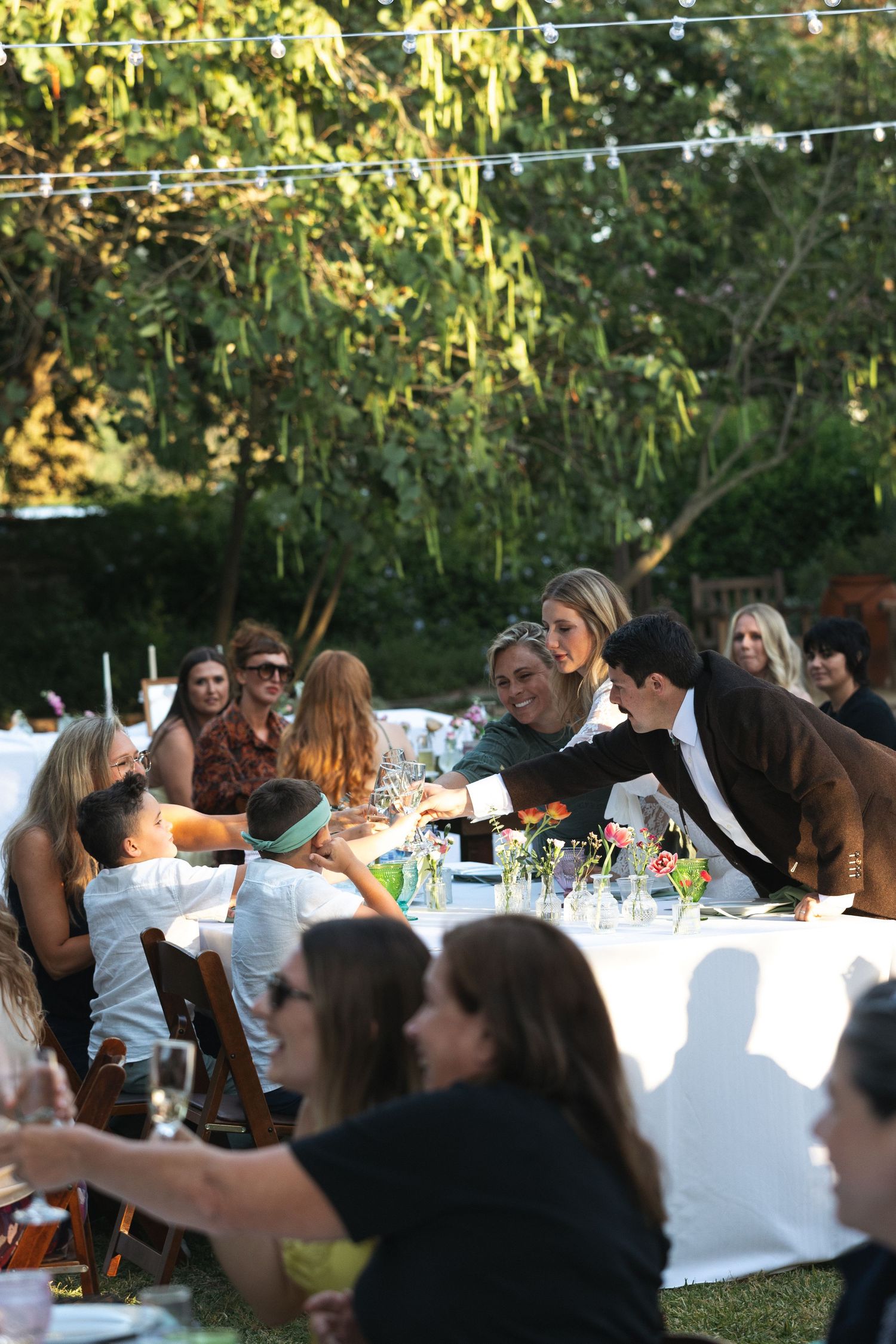 Guests gathered at an outdoor wedding reception dinner table, with a man pouring drinks amid lush greenery.