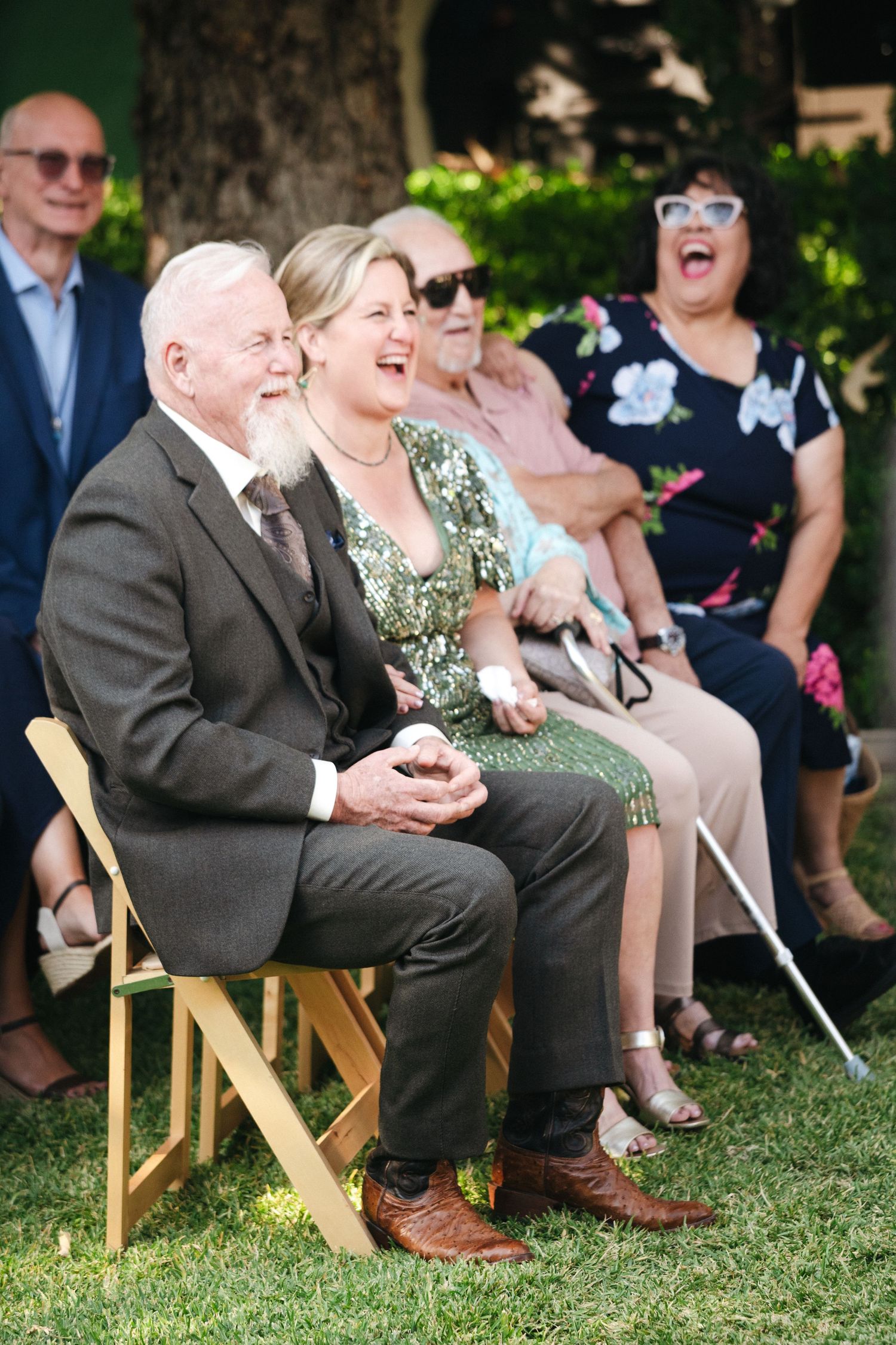 Guests laughing and smiling at an outdoor wedding ceremony, seated on wooden folding chairs on green grass.