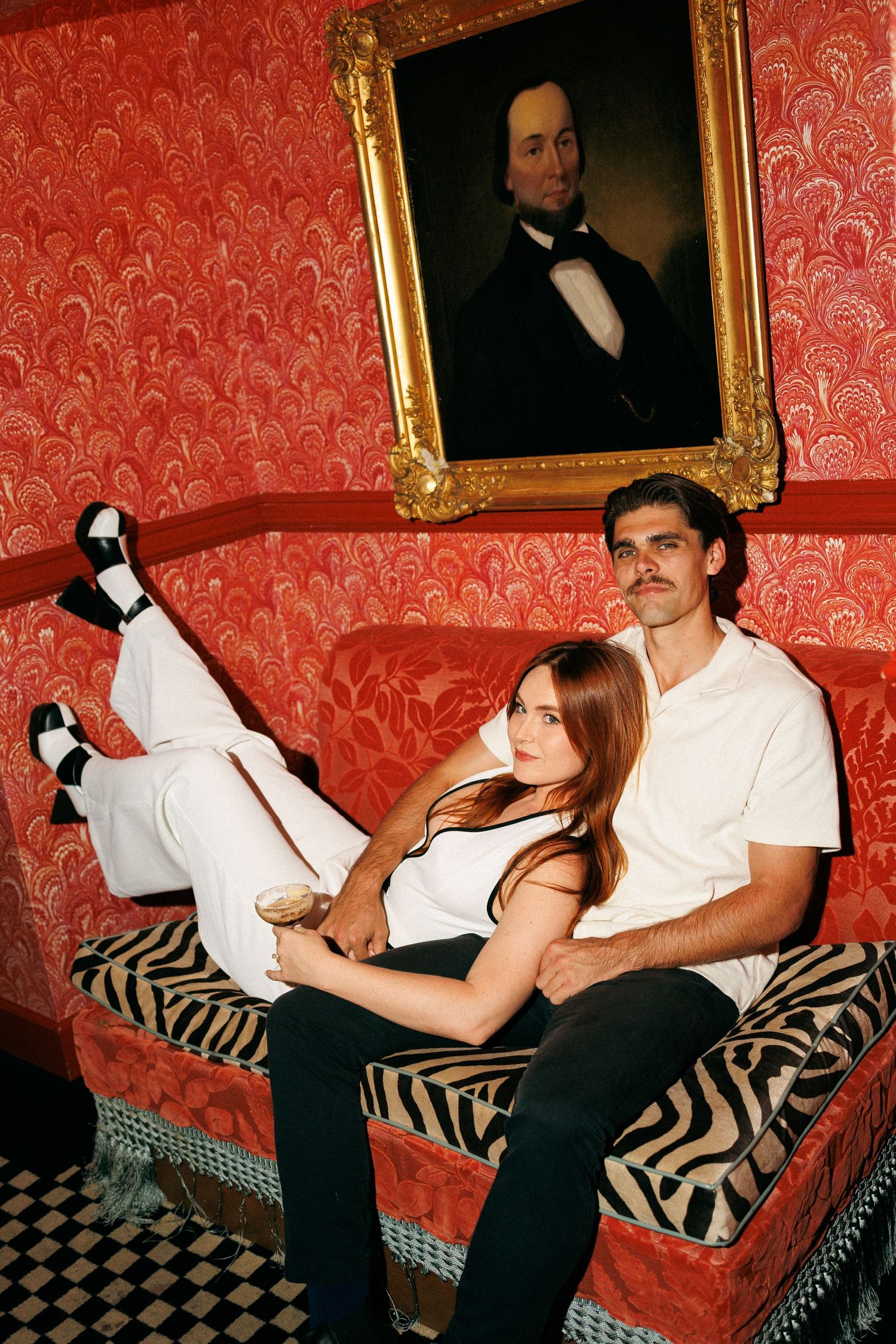 Couple relaxing on red velvet sofa beneath vintage portrait painting in ornate red wallpapered room.