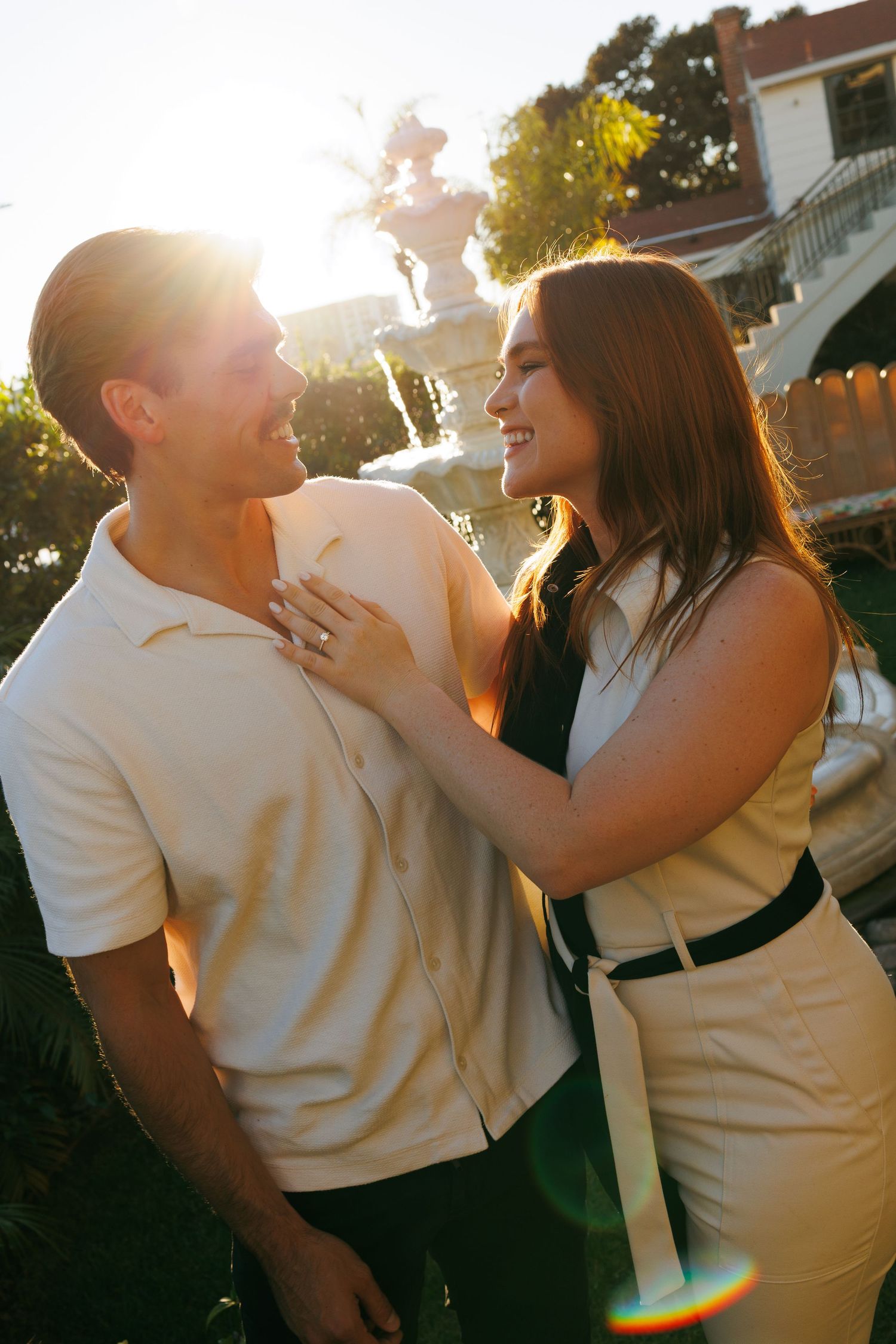 A couple smiles at each other in golden sunlight near a fountain, the woman placing her hand on the man's chest.