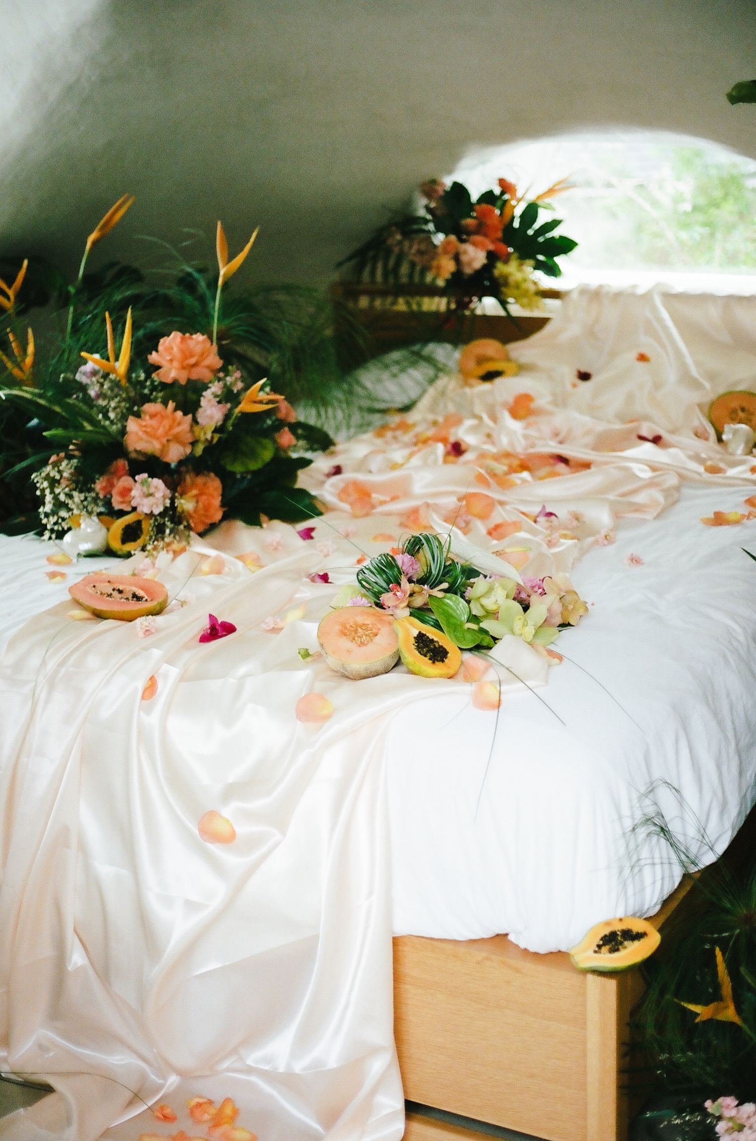 White bed decorated with tropical flowers, papaya, and scattered orange rose petals in a dome room.