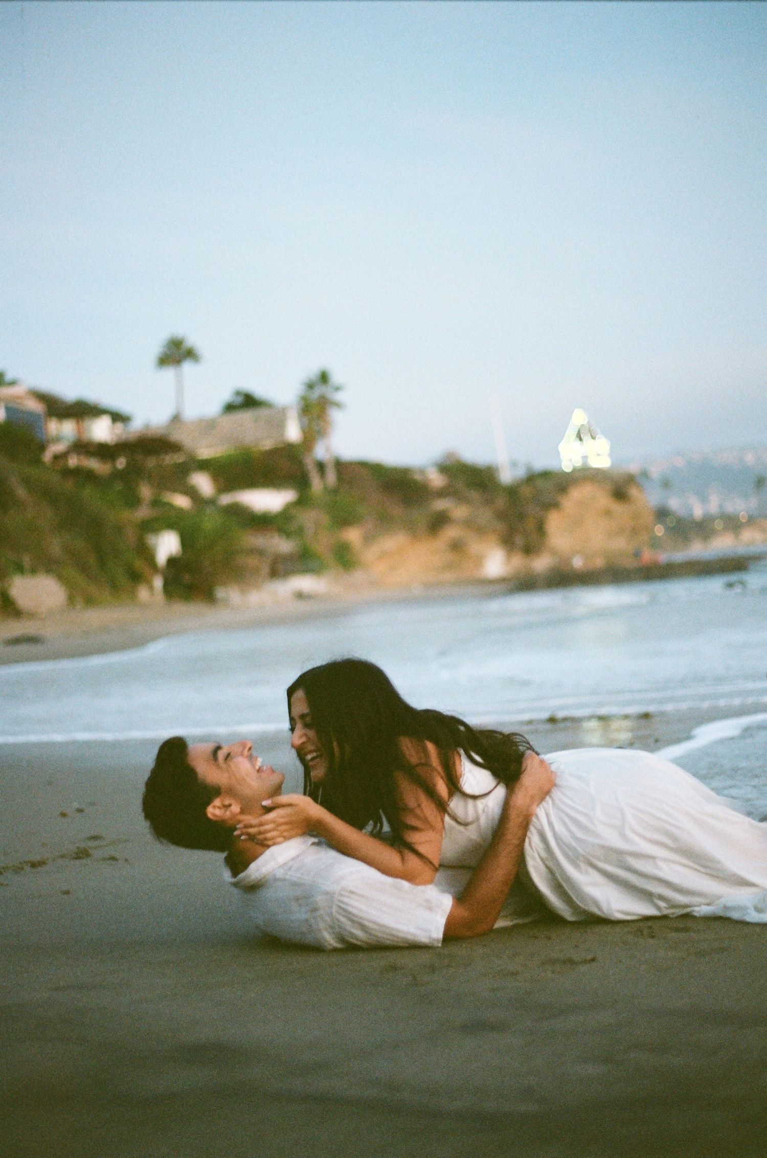 Romantic couple laughing and embracing while lying on a sandy beach at dusk near coastal cliffs.